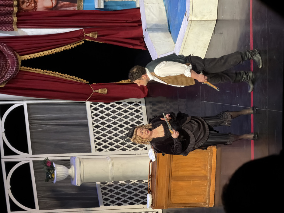 The image captures a scene from what appears to be a theatrical performance on a stage, viewed from the audience or wings. The image is rotated 90 degrees clockwise, so I will describe it as if it were correctly oriented.
Two actors are central to the frame. On the left, a man is dressed in historical attire, featuring a white collared shirt, a light brown or tan waistcoat, and dark trousers tucked into dark boots. He has short, dark, curly hair and is looking downwards and slightly to his left, with a focused expression. He is holding a small, ornate, dark-colored pistol or flintlock-style gun in his right hand, pointing it downwards. His posture suggests a moment of caution or suspense.
On the right, a woman is also in historical costume, wearing a dark, possibly black, velvet or heavy fabric dress or coat with a ruffled neckline and sleeves. Her outfit includes dark striped breeches or culottes and dark stockings or tights with dark shoes. She has blonde, curly hair styled in an updo and is wearing a dark, possibly feathered, small hat or hairpiece. She is looking to her left, towards the man, with a serious or concerned expression. She is holding a similar, ornate pistol or gun in her left hand, also pointed downwards, near her waist. She stands near a wooden piece of furniture that resembles a desk or cabinet, which is a medium brown color.
The stage set behind them features several elements. To the far left, a large red curtain with gold trim and tassels is partially drawn, revealing a dark, empty space behind it, suggesting an entrance or exit point. Above this curtain, part of what looks like a painted backdrop or flat showing a classical or mythological figure (possibly a cherub or putto) with an elaborate hairstyle is visible.
To the right of the red curtain, there's a white stage flat designed to resemble architectural elements. It includes sections with white lattice work and panels that mimic windows with sheer, white translucent curtains behind them. In the lower left portion of the flat, a white classical column is visible, topped with a white urn containing a small arrangement of colorful artificial flowers (pink, yellow, green).
The stage floor is dark, possibly black, with a distinct red line running horizontally across it in the midground, suggesting a stage boundary or marking. The lighting appears to be typical stage lighting, illuminating the actors and set pieces. The overall impression is a dramatic moment within a period play or opera. No discernible text is visible in the image.