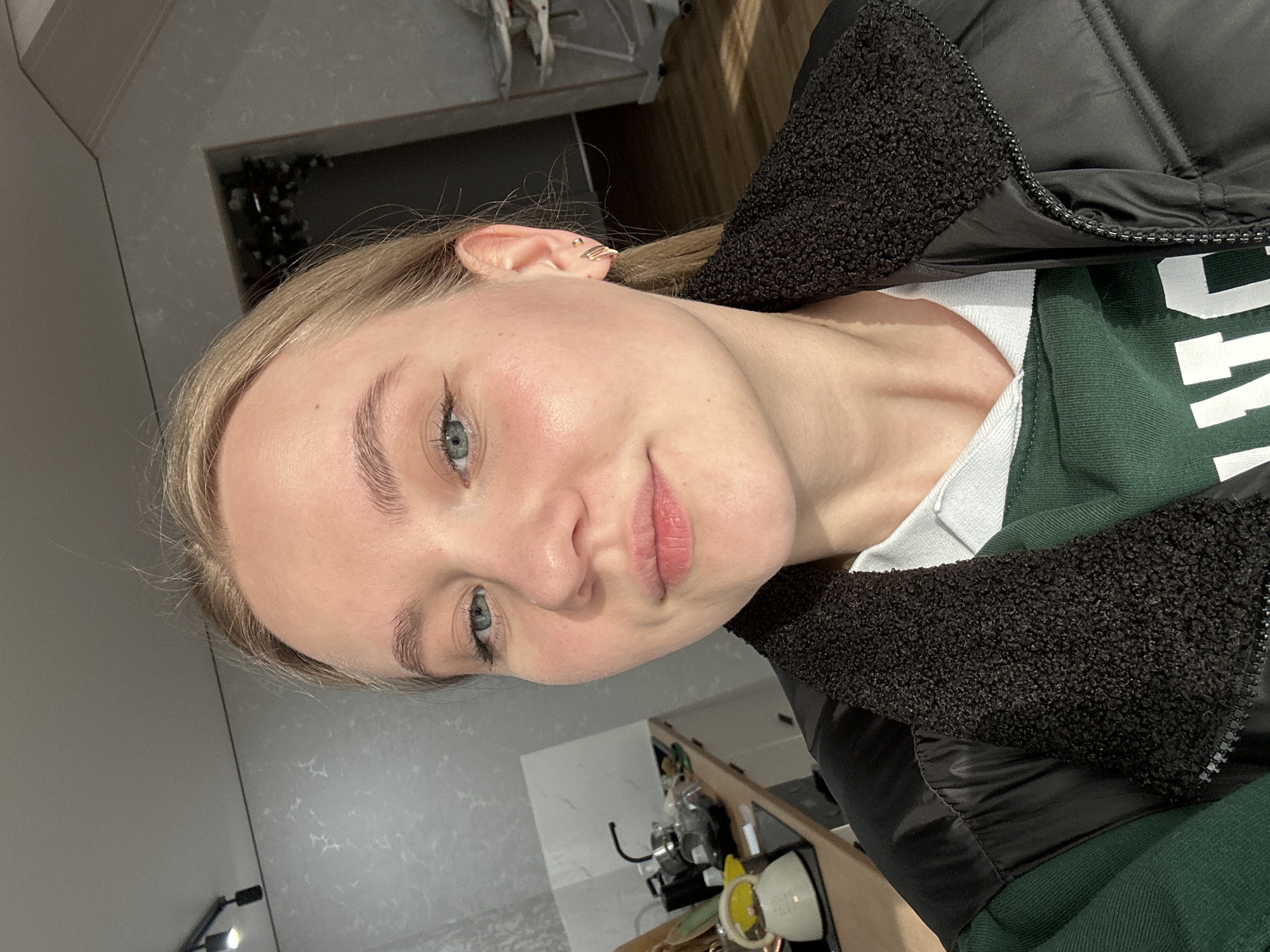 An indoor, close-up portrait shows a young woman with fair skin, light blonde hair, and blue-green eyes, looking directly at the camera with a gentle, slightly smiling expression. She has well-groomed eyebrows and subtle winged eyeliner. A small, delicate gold-colored ear cuff or piercing is visible on her right ear.
She is wearing a black puffer jacket with a textured, dark, possibly fleece or sherpa, lining visible along the collar and inside the sleeve cuffs. Underneath the jacket, a white t-shirt and a dark green sweatshirt or sweater are visible.
The background suggests a casual indoor setting, possibly a home in Chaikovsky, Russia. To the upper right, a dimly lit area reveals some indistinct dark objects and possibly decorative elements like Christmas lights or garland. To the lower right, part of a counter or table with some household items, possibly kitchen appliances, is visible. The lighting appears to be natural, coming from the left, illuminating her face. No discernible text is visible in the image.