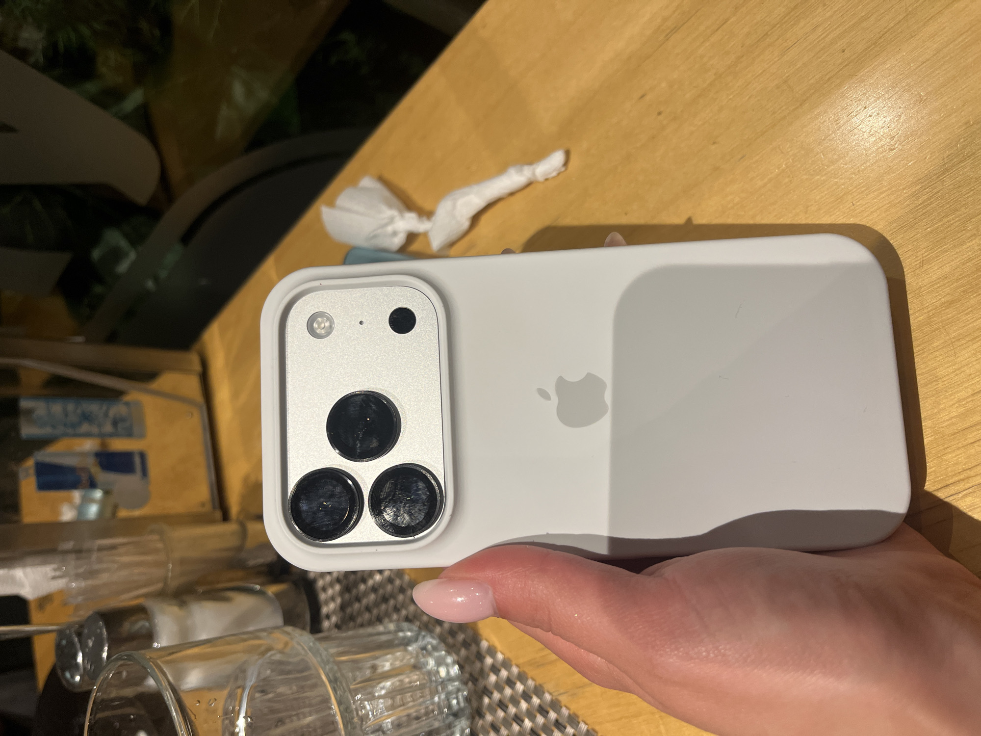 A person's hand, with lightly polished pink fingernails, holds a white iPhone case with a triple-lens camera array. The case is positioned on a light brown wooden table, with a patterned placemat visible on the left side of the frame. In the background, out of focus, are salt and pepper shakers, a glass, and what appear to be beverage containers on a shelf. The overall setting suggests an indoor, possibly casual dining environment. No specific location in Plovdiv, Bulgaria can be determined from the provided image.