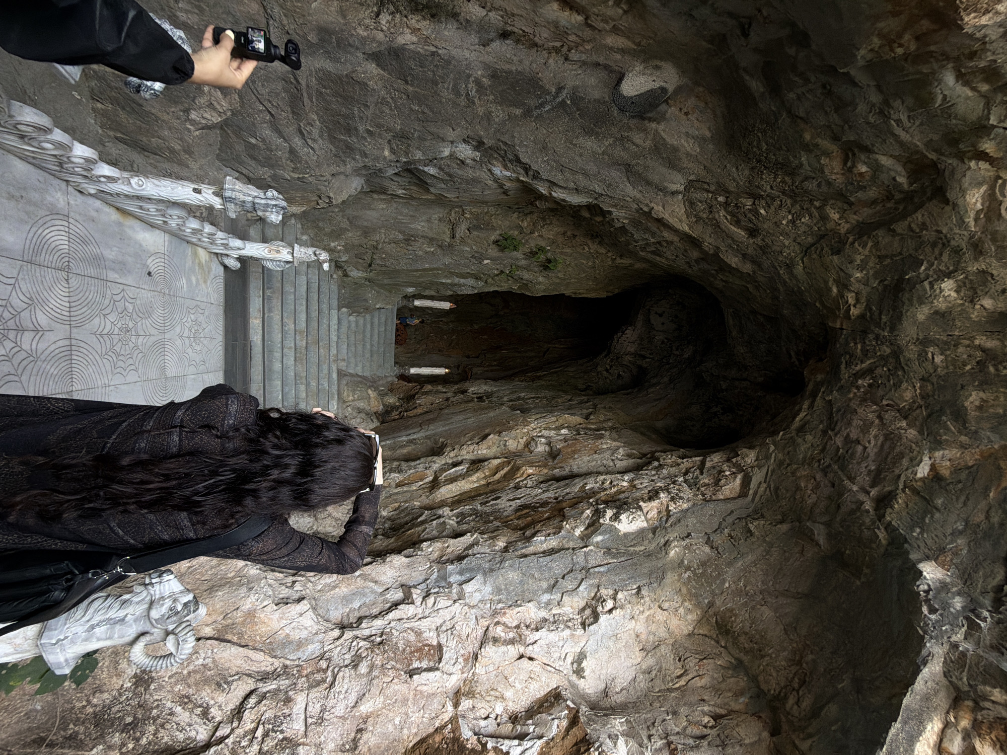 Two individuals stand at the edge of a stone staircase leading into a cave-like opening in Đà Nẵng, Vietnam. One person is positioned to photograph the cave interior with a handheld device, while the second person uses a camera. The architecture features carved stone railings depicting animals and decorative patterns. The cave is dark with light sources visible in the distance. The surrounding environment is characterized by exposed rock formations.