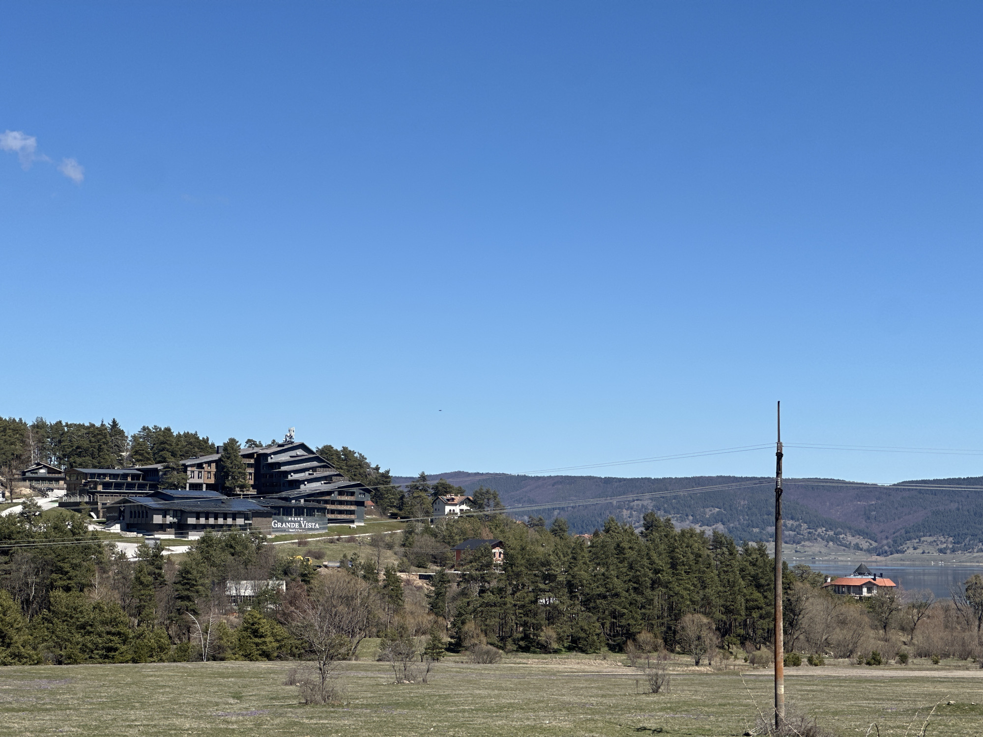 The image depicts a landscape in Цигов чарк, Bulgaria, under a clear blue sky with a few wispy clouds. The Grande Vista resort is visible on a hillside, nestled among trees. In the foreground is a grassy field. A power pole with wires extends vertically from the field. Beyond the resort and trees, there are hills and a glimpse of a lake. A house with a red roof is visible on the far right near the water.
