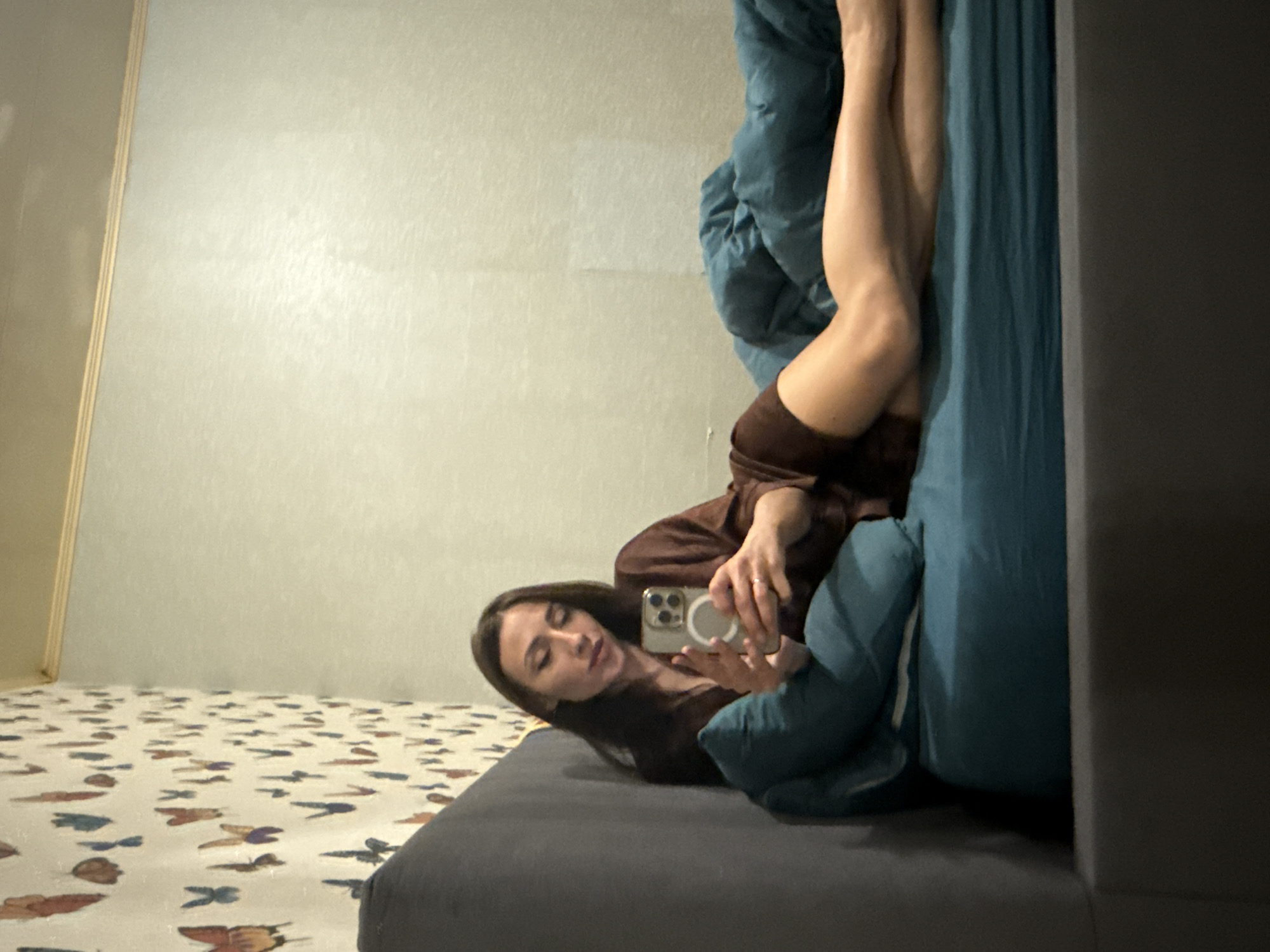 The image captures a young woman lying in a bedroom or personal living space in Saint Petersburg, Russia. She has long dark hair and is dressed in a brown top or robe. Her head rests on a dark gray mattress, and she holds a smartphone with a prominent camera module and a white circular attachment on its back, seemingly taking a selfie or interacting with the device. Her legs are extended upwards, resting against blue bedding.
The room features a light-colored, subtly textured wall, with a vertical wooden-toned trim visible on the left. The floor is covered by a light-colored rug or carpet adorned with numerous small, colorful patterns resembling butterflies or abstract shapes in hues of brown, blue, and reddish-orange. The lighting is soft and even, suggesting an indoor setting without direct sunlight. The woman's expression is neutral and focused on her phone. There is no visible text in the image.