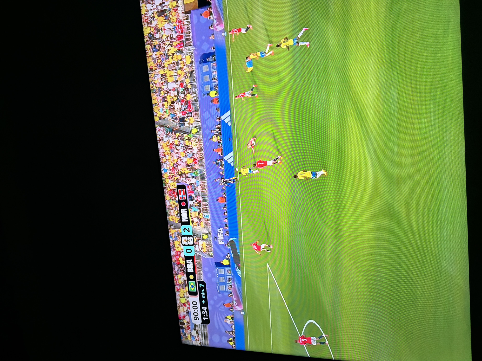 The image shows a soccer match being broadcast on a television screen. The score indicates that BRA is leading NOR 2-0, with the game in the 90th minute plus 1 minute and 34 seconds of added time. The field is green, and players in red and yellow/blue uniforms are visible on the field. The stands are filled with spectators wearing a variety of colored clothing. The television screen is angled, suggesting it is being viewed in a room with limited lighting. The image does not provide any information about the city of Ognianovo, Bulgaria, or any context linking it to this scene.