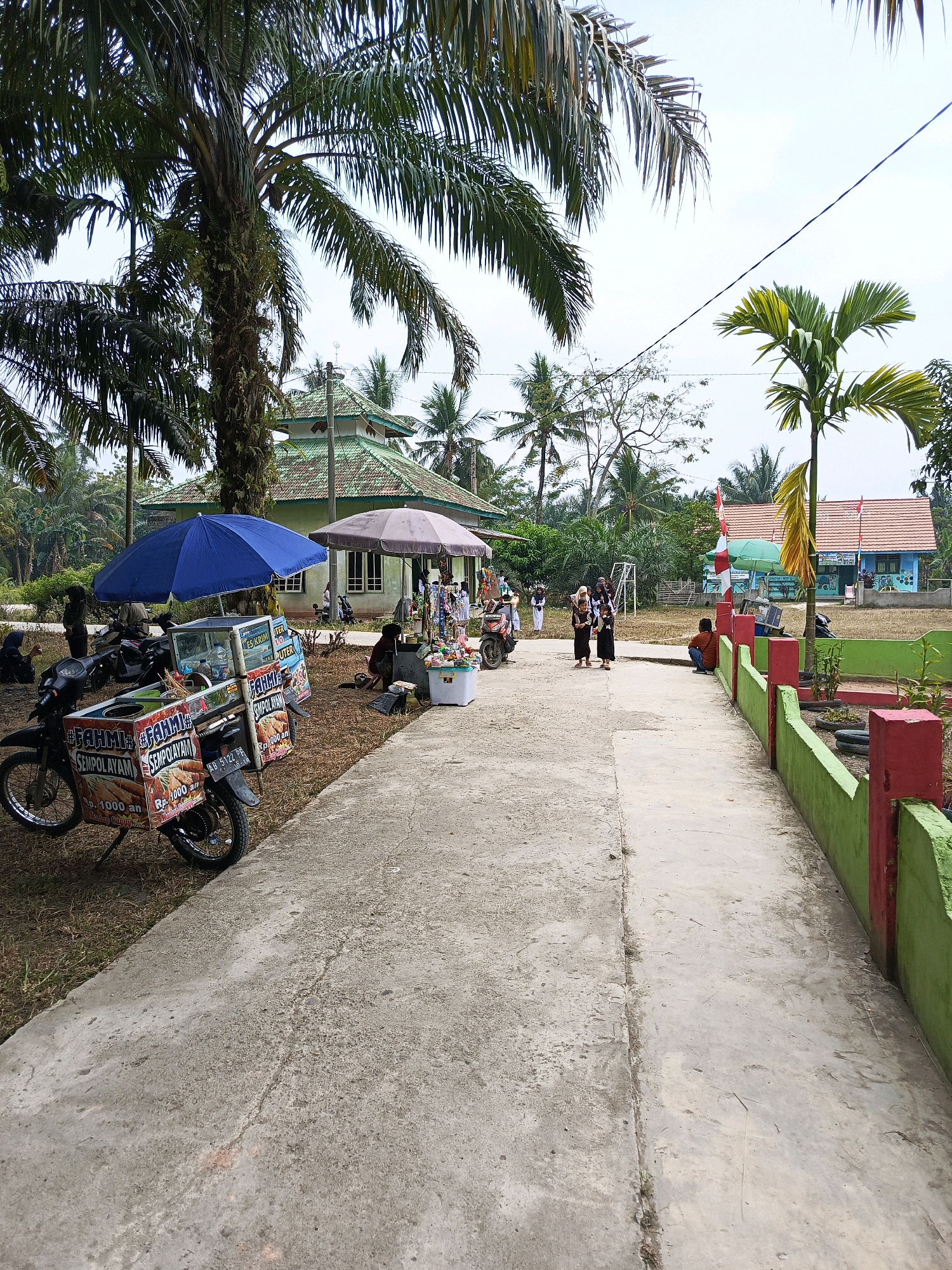 In the city of Megang Sakti V, Indonesia, a concrete pathway leads through a grassy area dotted with palm trees. On the left side of the path, a motorcycle with a food cart attached is parked under a blue umbrella. The cart displays signage for "FAHMI SEMPOL AYAM" and "ES KRIM," indicating it sells fried chicken skewers and ice cream. Several people are gathered around the food cart and in the vicinity. Further down the path, a large, weathered umbrella shades another area where a few individuals are visible. In the background, a modest green-roofed building, possibly a mosque or a community hall, stands amidst the trees. To the right of the pathway, a low green and red fence encloses a garden area. Beyond this, a brightly colored blue building with an orange roof, likely a school, is visible, featuring a red and white Indonesian flag flying in front. The scene appears to be a lively outdoor gathering, possibly a local market or event, under a clear sky.