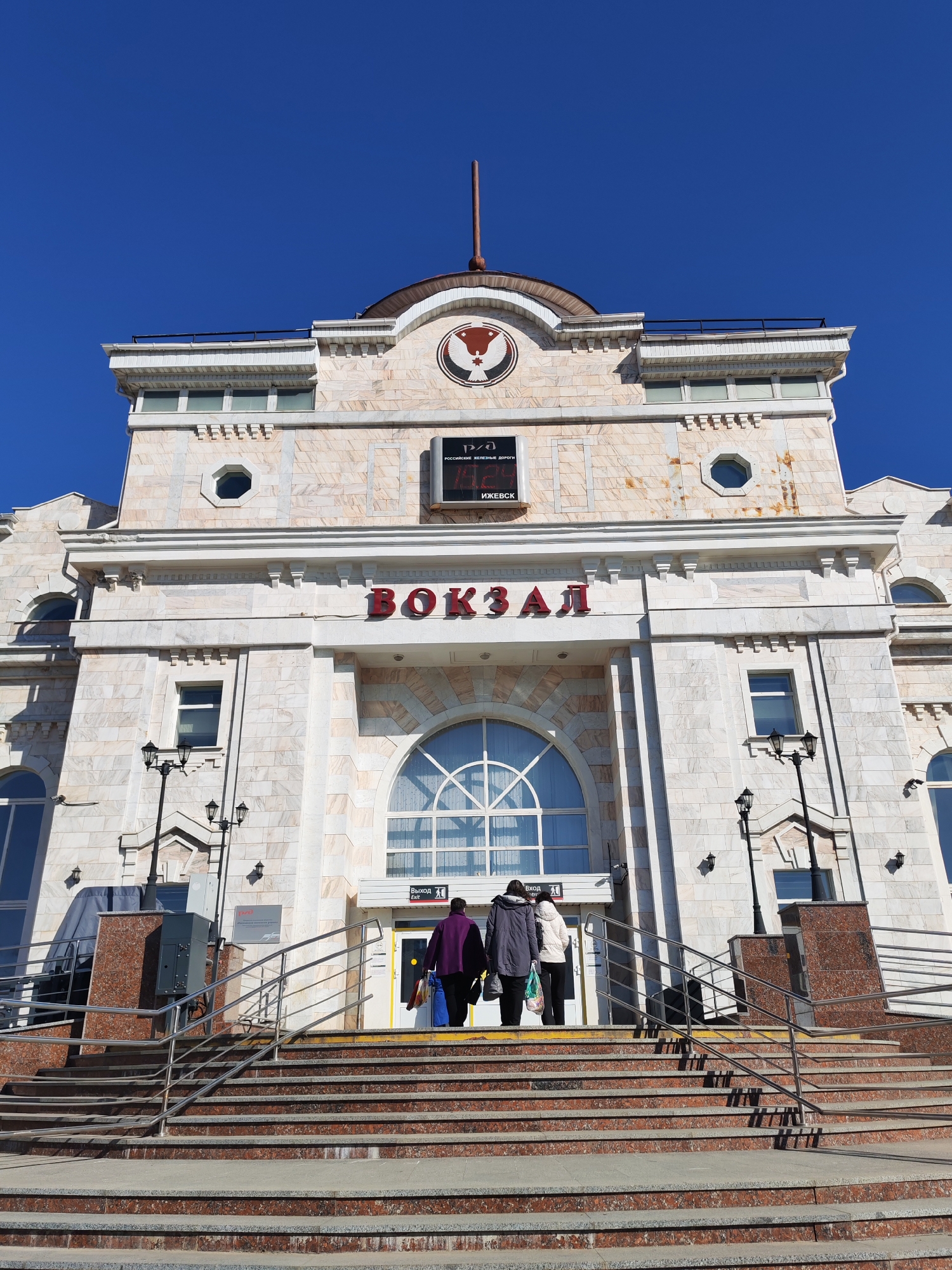 The image presents the grand facade of a train station in Izhevsk, Russia, bathed in bright sunlight under a clear, deep blue sky. The building exhibits a neoclassical architectural style, constructed from light-colored, possibly marble or granite, stone with decorative cornices, pilasters, and a prominent central pediment. A flagpole rises from the top of the structure, and a circular emblem featuring a stylized bird (the Russian Railways logo) is visible on the pediment.
Above the main entrance, large red Cyrillic letters clearly spell "ВОКЗАЛ" (VOKZAL), meaning "Station." Below the pediment, a sign further identifies the location with "РОССИЙСКИЕ ЖЕЛЕЗНЫЕ ДОРОГИ" (Russian Railways) and "ИЖЕВСК" (Izhevsk). The entrance is accessed via a wide set of reddish-brown and gray stone steps, equipped with stainless steel handrails.
Three individuals, seen from behind, are walking up these steps towards the station's main doors. They appear to be travelers, carrying bags and dressed in coats. A sign near the entrance doors reads "Выход" (Exit). The building features various window designs, including a large arched window above the main entrance, and smaller octagonal and rectangular windows on the upper levels. Two ornate black lampposts flank the stairs, adding to the classic aesthetic of the station. The strong sunlight casts distinct shadows, highlighting the architectural details and the activity of the people.