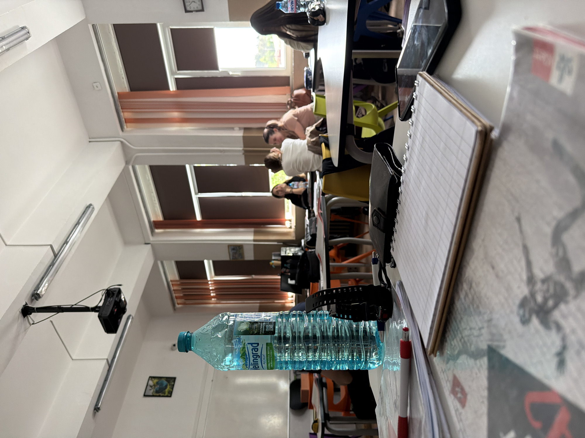 The image shows the interior of a classroom, likely in Panagyurishte, Bulgaria, as indicated by the "Elingrad" water bottle on the table. The classroom has multiple rows of desks where students are seated. The students are facing the front of the classroom. Windows are visible on one side of the room. Desks and books are prominent features in the foreground. A security camera is mounted on the wall.