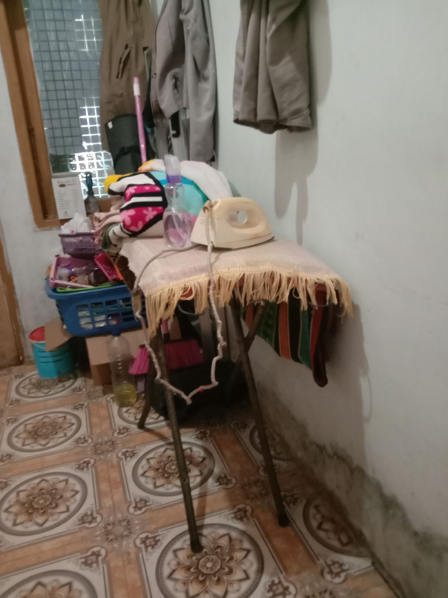The image depicts an ironing board with a cream-colored iron resting on top, situated in a dimly lit room with tiled flooring. A patterned cloth with a fringe hangs from the ironing board. To the left of the ironing board, a blue plastic basket contains various items, including what appear to be cleaning supplies. A red bucket and a clear plastic bottle with yellow liquid are also visible on the floor. In the background, clothes are hanging from a rack near a window with a grid pattern. The walls of the room appear to be a pale color. The overall scene suggests a domestic setting, possibly a laundry area, located in Kwandang, Indonesia.