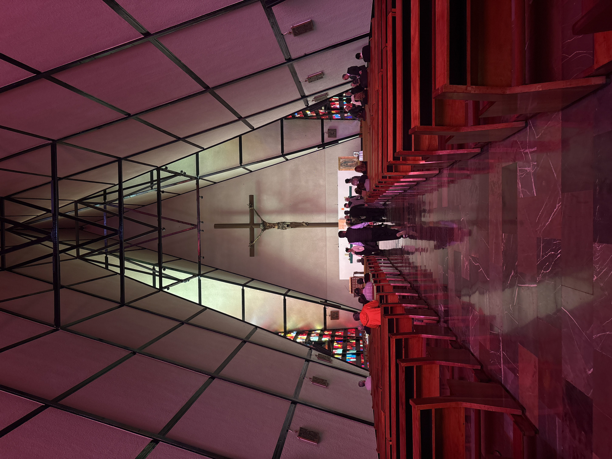 This is an interior shot of a modern church with a distinctive A-frame roof structure. The walls are paneled with what appears to be a light-colored material framed by dark metal beams, creating a series of triangular sections. The ceiling tapers to a peak, revealing a black metal truss system. Stained glass windows, illuminated with vibrant colors, are visible on the left and right sides, further up the walls, and also as a triangular array in the apse.
At the far end of the church, a wooden crucifix is mounted above a white altar area. In front of the altar, a small painting of the Virgin of Guadalupe is displayed. The church is furnished with rows of dark wooden pews. The floor is made of polished marble tiles in shades of dark red and black, reflecting the ambient light.
Several individuals are present in the church. Some are seated in the pews, while others are standing and walking. In the foreground, the rows of empty pews are prominent. Towards the center of the image, a group of people are walking down the aisle. A man in a red jacket is seated in the middle of a pew on the left.
The lighting in the church is predominantly in shades of pink and red, casting a warm and dramatic glow throughout the space. The overall impression is one of a contemporary architectural design for a place of worship, with a solemn yet vibrant atmosphere. The church is likely located in Mexico City, Mexico, given the presence of the Virgin of Guadalupe painting and the architectural style often found in the region.