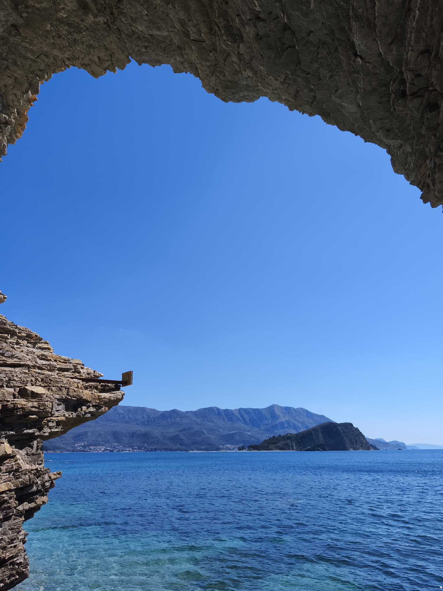 The image depicts a view through a rock formation, possibly a cave entrance, overlooking a body of water. The sky is a clear, vibrant blue. In the distance, there are mountains and a small island. The water is a deep blue color with visible ripples and some areas of clearer, turquoise water near the rock formation. The rock formation itself is layered with different shades of brown and gray. On the left side, a protruding rock has what appears to be a small, rectangular metal fixture attached to it.