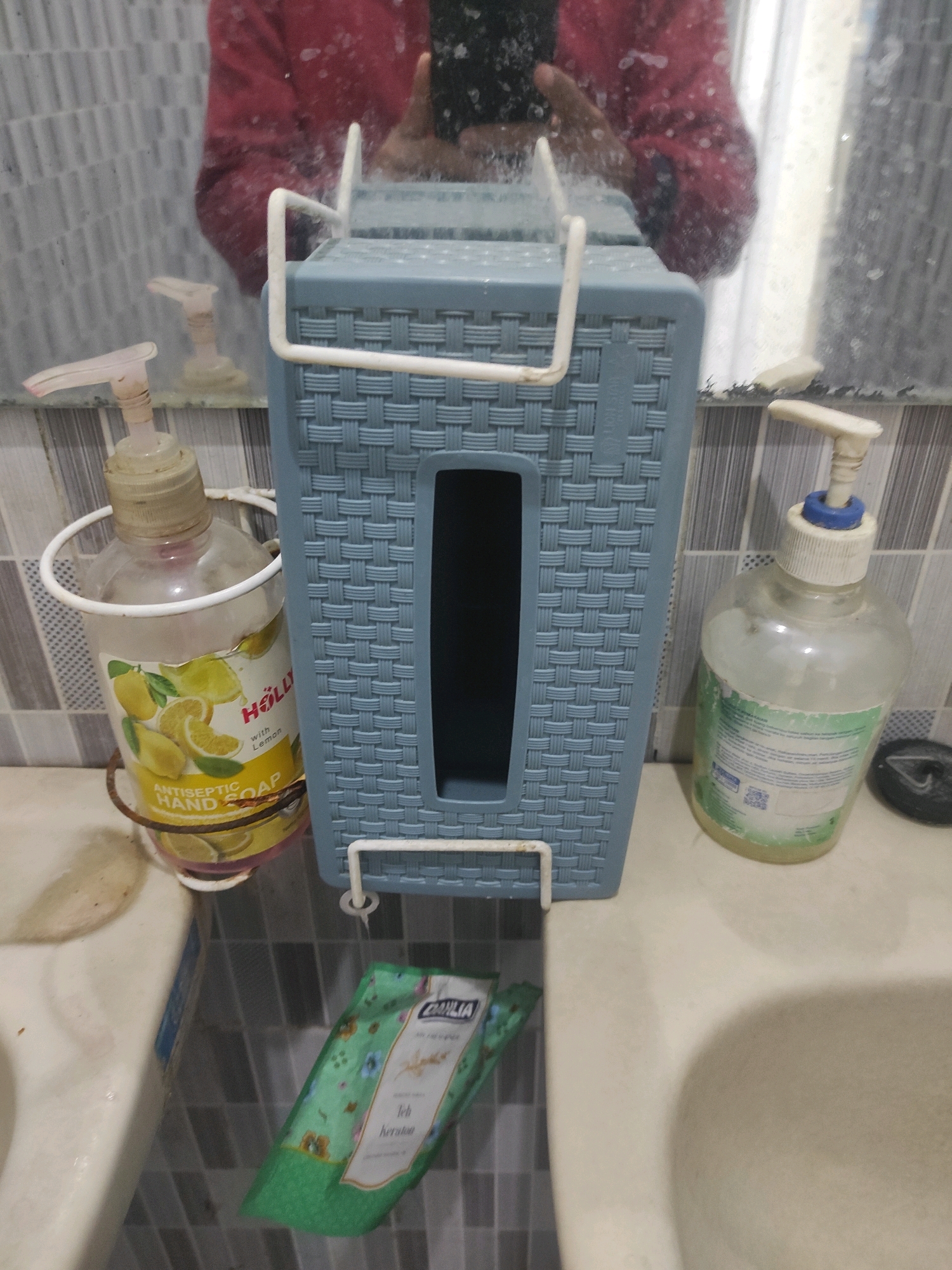The image depicts a sink area in a bathroom. In the center of the image, a blue plastic dispenser with a woven texture is mounted. To the left of the dispenser, a bottle of "HOLLY Antiseptic Hand Soap with Lemon" is hanging in a wire holder. To the right of the dispenser, another bottle of liquid soap with a blue cap is placed on the counter. Below the blue dispenser, a green packet with floral patterns and text that appears to be "Teh Kera" is lying on the sink's tiled surface. The sink itself is made of white ceramic and is part of a countertop. The background shows a tiled wall and a mirror reflecting a person wearing a red garment. The presence of hand soap and the general arrangement suggest a functional bathroom sink area. This scene is located in Bojonegoro, Indonesia.