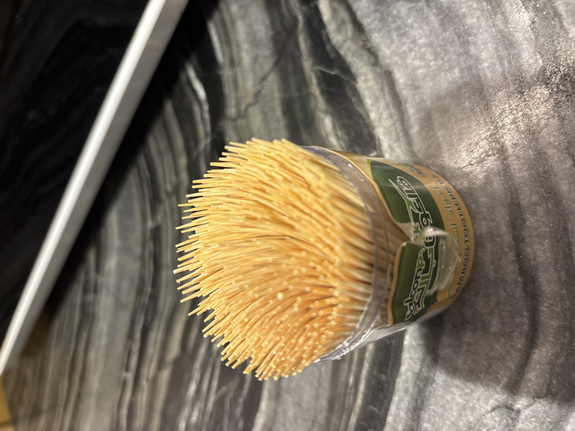A clear plastic container filled with a bundle of natural wood colored toothpicks sits on a dark grey, marbled countertop. The container has a green and yellow label with Arabic script and the words "BAMBOO TOOTH PICKS" printed in English. A portion of a white, rectangular object is visible in the upper part of the image, casting a subtle shadow. The background is dark and out of focus, with streaks of lighter grey. The scene appears to be indoors, likely in a kitchen or dining area, possibly in Dubai, United Arab Emirates, given the presence of Arabic script on the label.