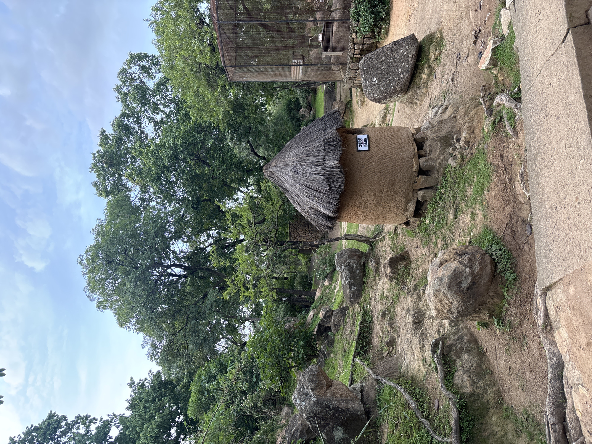 The image shows an outdoor scene with a small, round hut with a thatched roof. The hut is made of mud and is elevated on stones. A sign on the hut reads "FERE BARN". To the right of the hut is a large, wire-mesh enclosure, suggesting it might be a zoo or wildlife sanctuary. The environment is natural, with trees, rocks, and grass visible. The ground is sandy and uneven. The sky is cloudy. This scene is located in the city of Jos, Nigeria, likely at the zoo or a similar facility. There are no people visible in the image.