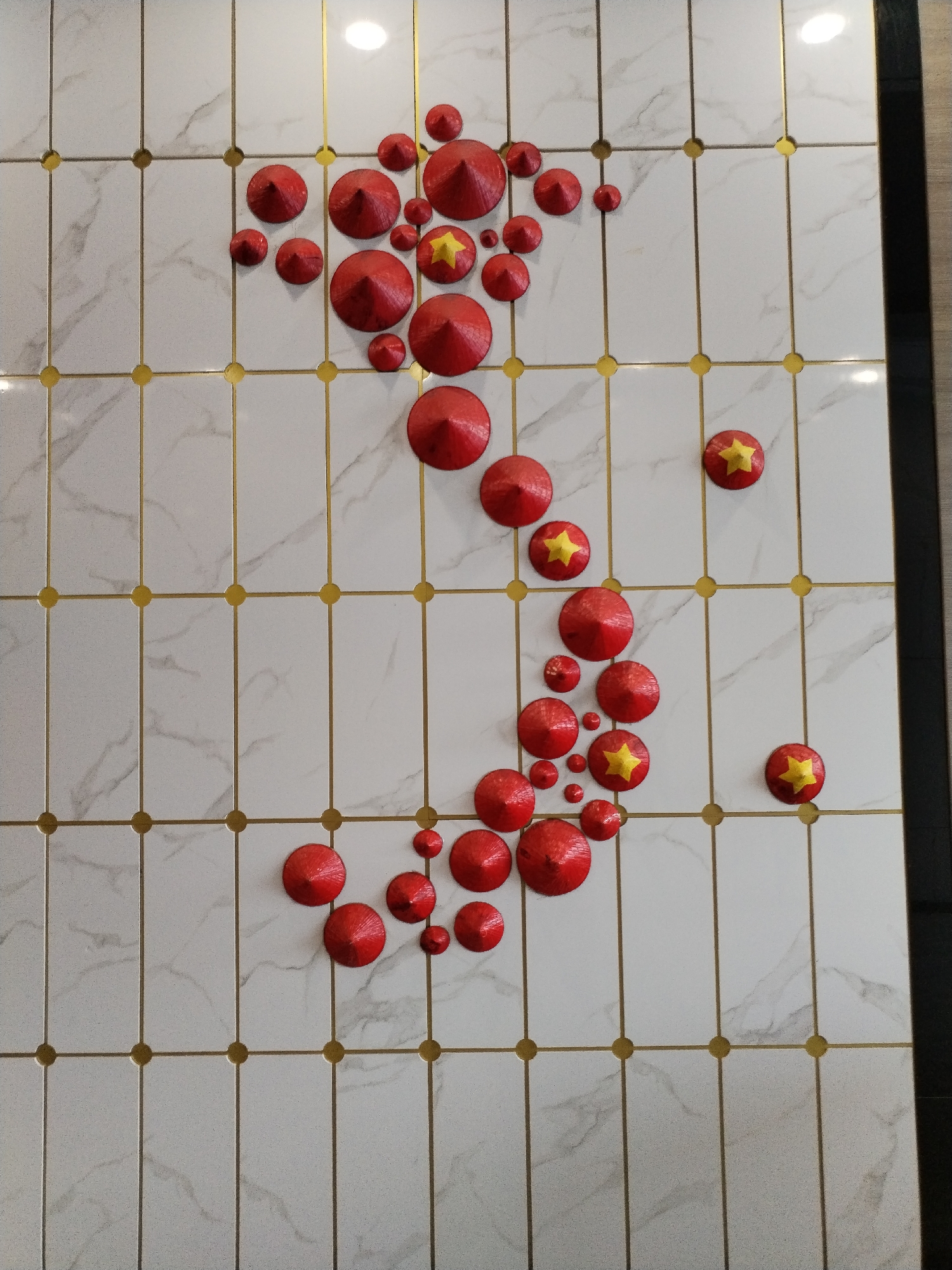 The image depicts a wall decoration in what appears to be a public space, possibly a restaurant or hotel, in Nha Trang Ward, Vietnam. The wall is covered with white marble-patterned tiles, separated by gold grout lines that form a grid. Scattered across the wall are numerous red conical hats, commonly known as "nón lá," which are traditional Vietnamese hats. Some of these hats are plain red, while others are adorned with a yellow star, resembling the Vietnamese flag. The hats are arranged in a pattern that loosely outlines the shape of Vietnam. The lighting suggests an indoor environment, with soft illumination reflecting off the glossy tile surface. There are no people visible in the image.