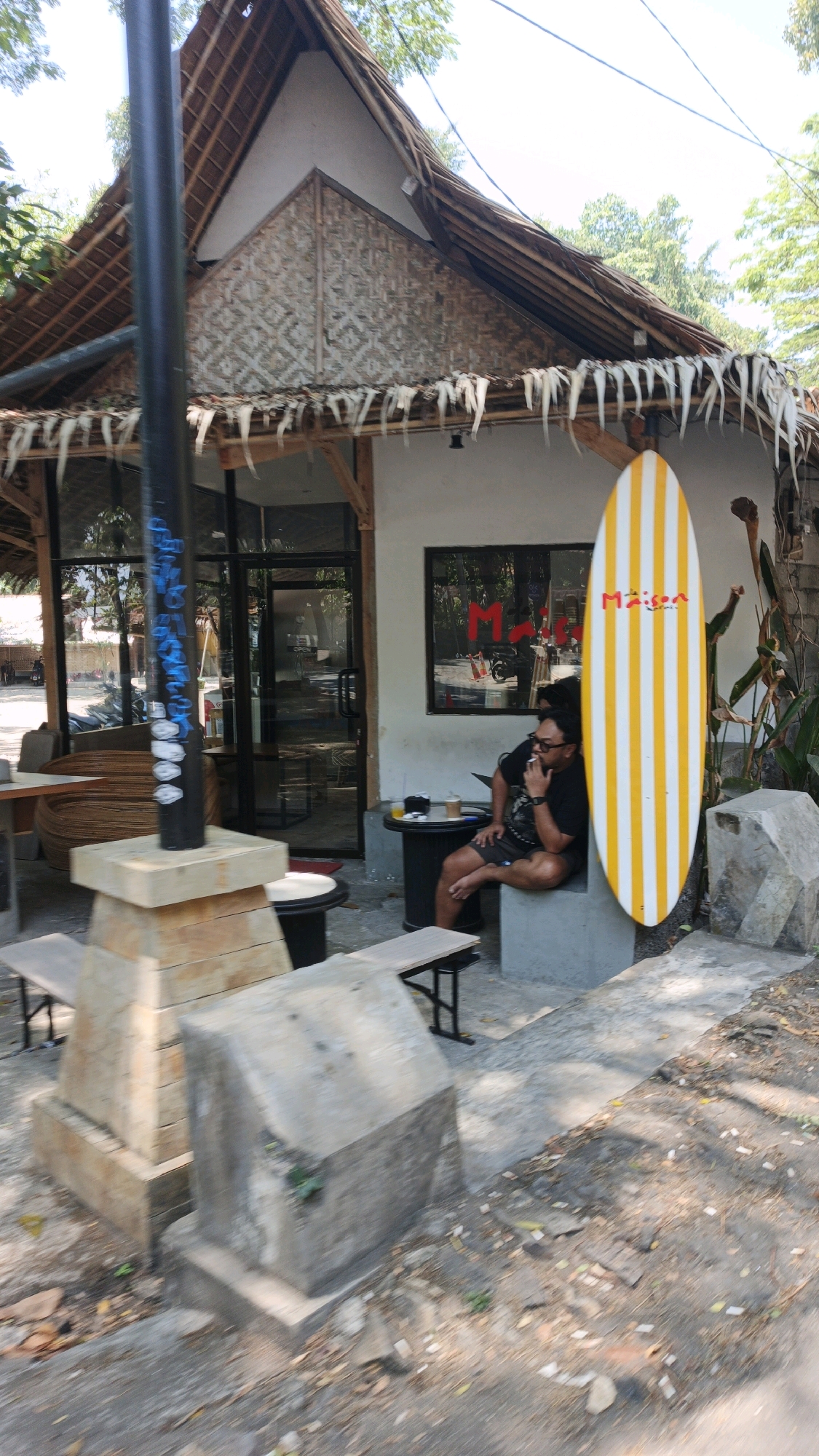 This image depicts the exterior of a cafe or shop in Desa Batukaras, Indonesia, with a distinct tropical, possibly beachside, ambiance. The building features a thatched roof with hanging decorations and a wall decorated with woven bamboo. In front of the building, a man is seated on a concrete block, smoking a cigarette and looking to his left. He is wearing a black t-shirt and shorts. To his right, a surfboard with yellow and white vertical stripes stands upright against the building. A round black table is positioned next to the man, holding two cups of beverages. Several concrete structures and wooden benches are visible in the foreground, suggesting an outdoor seating area. The ground is paved with concrete and scattered with fallen leaves and debris. The background shows trees and foliage, indicative of a warm climate. The overall scene suggests a casual, relaxed setting.