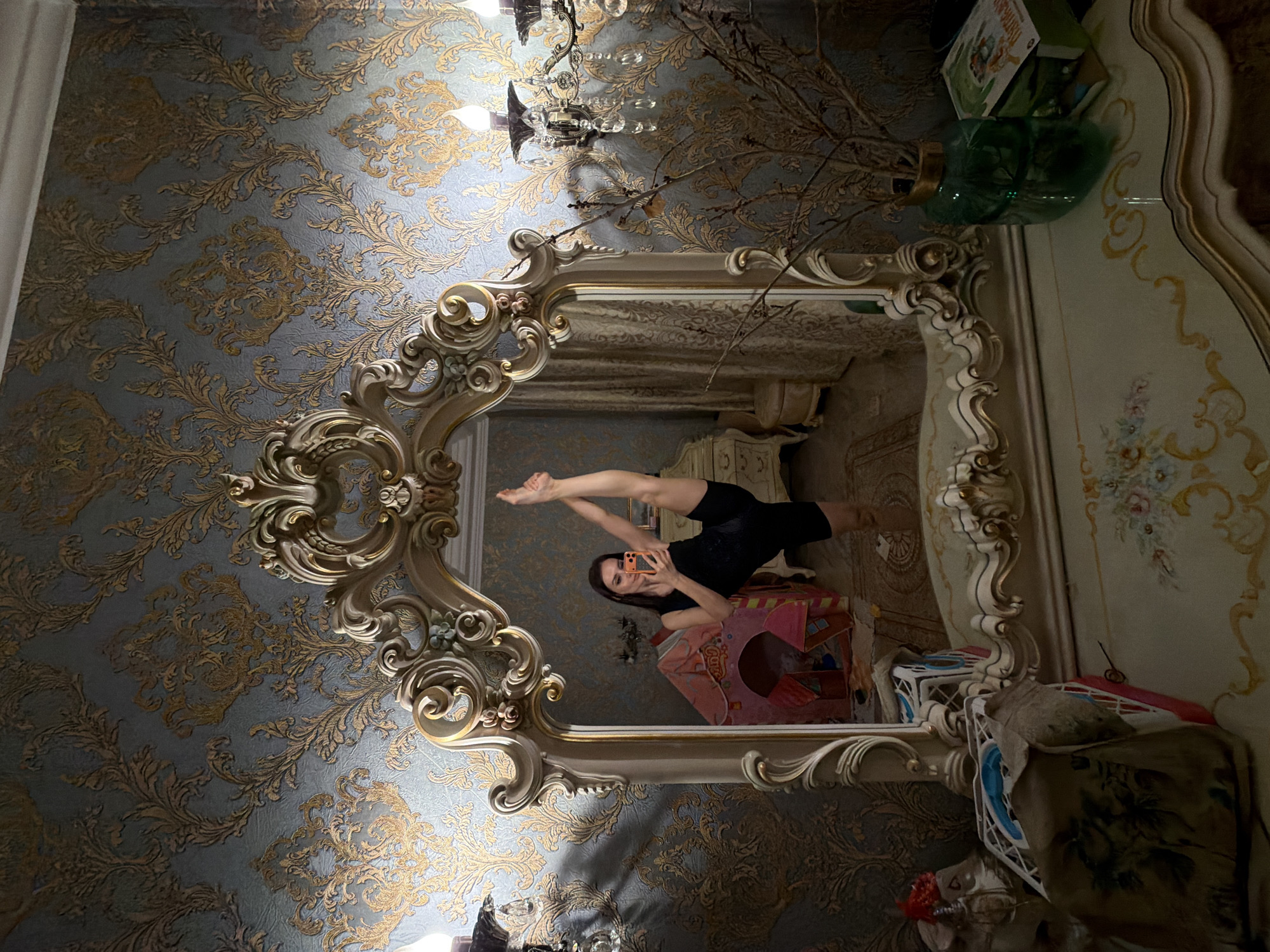 A woman is captured in a mirror's reflection, performing a high leg split. She is positioned in the center of the frame, her right leg extended vertically upwards, while her left leg remains bent. She is holding a smartphone in her right hand, presumably to take the photo. The woman is wearing a black tank top and black shorts. She stands in a room with ornate wallpaper featuring a gold damask pattern on a blue background. The mirror itself is elaborate, framed by a gilded, carved baroque style. To the left and right of the mirror are wall-mounted light fixtures with crystal accents. In the background, reflected in the mirror, is a child's pink play tent and part of a dresser. The foreground, below the mirror, shows the surface of a vanity table with some decorative items.
