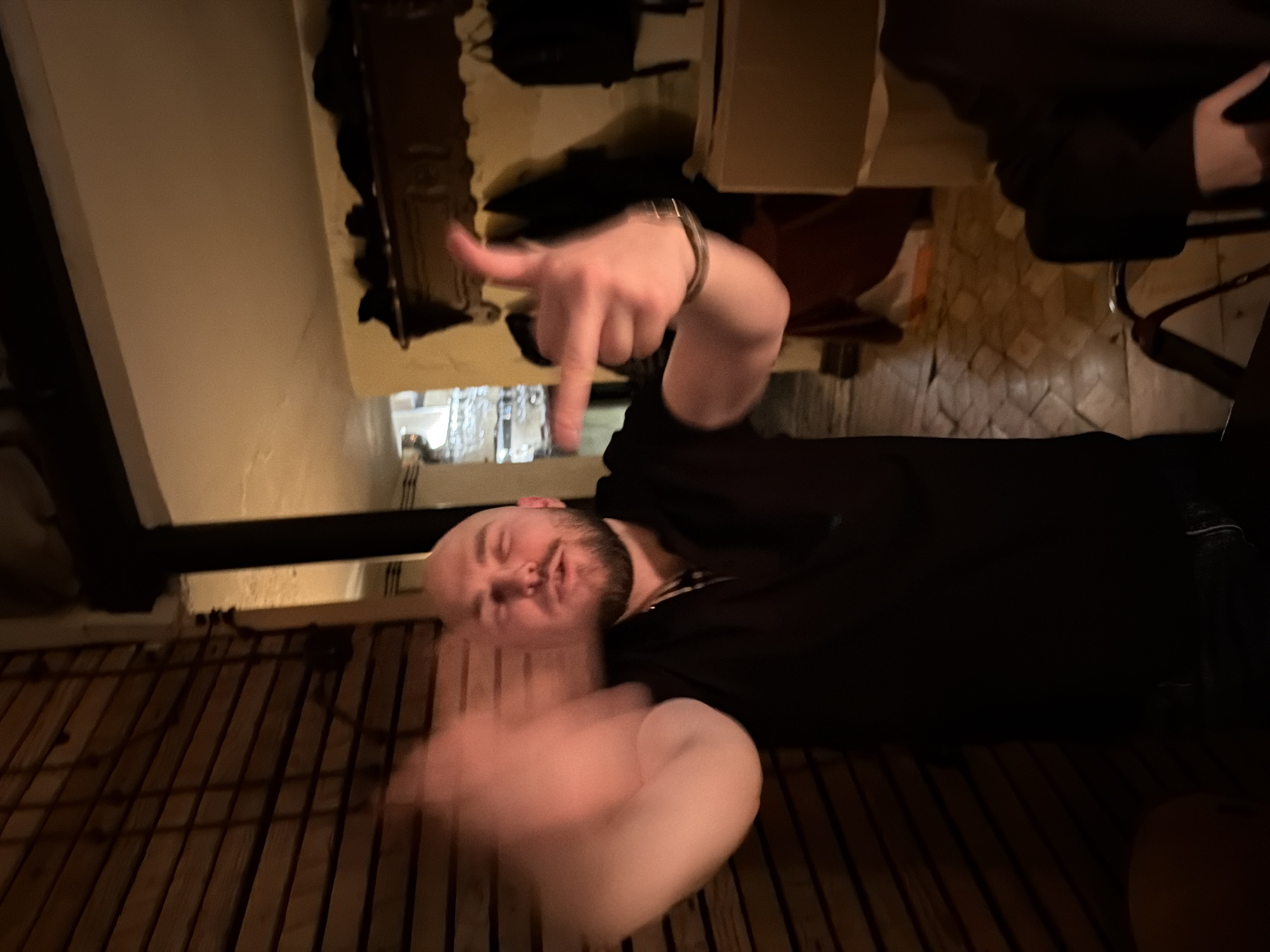 The image depicts a dimly lit indoor scene in Tbilisi, Georgia, likely an establishment such as a bar or restaurant. A bald adult male with a beard, wearing a dark polo shirt, stands centrally. His left hand is blurred due to motion, while his right hand, adorned with a metallic watch, forms a finger-gun gesture pointed towards the background. His eyes are partially closed or squinting, and his mouth is slightly open. To the viewer's left, a wooden slatted wall is visible. To the viewer's right and background, an open doorway reveals an adjacent room with light-colored walls, a dark wooden mantelpiece topped with dark items, and a further opening leading to an area with hanging glassware. The floor in the background appears to be tiled with a muted, patterned design. A brown cardboard box and the arm/legs of other individuals are partially visible in the lower right foreground. The specific location within Tbilisi cannot be identified from the provided visual information.