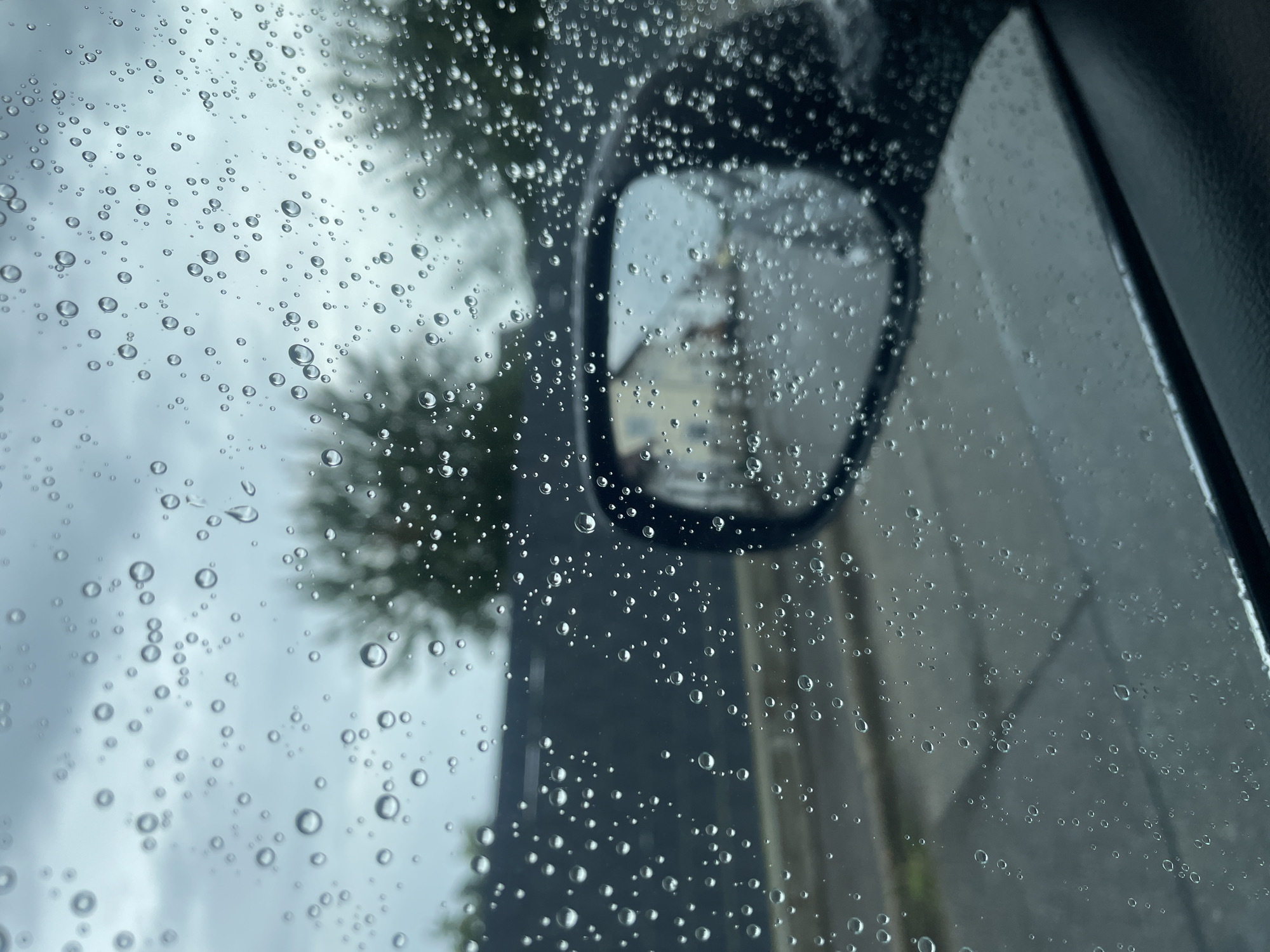 This is a photograph taken from inside a car looking through a rain-streaked window. Water droplets of varying sizes are scattered across the glass, obscuring the view of the outside. In the mid-ground, a dark-colored car with a side-view mirror is visible. The mirror reflects a blurry image of houses and a street. Beyond the car, trees and a building are visible through the rain. The sky appears overcast and cloudy. The photograph is likely taken in Geiselhöring, Germany, given the context, but no specific landmarks or identifying features in the image confirm this location.