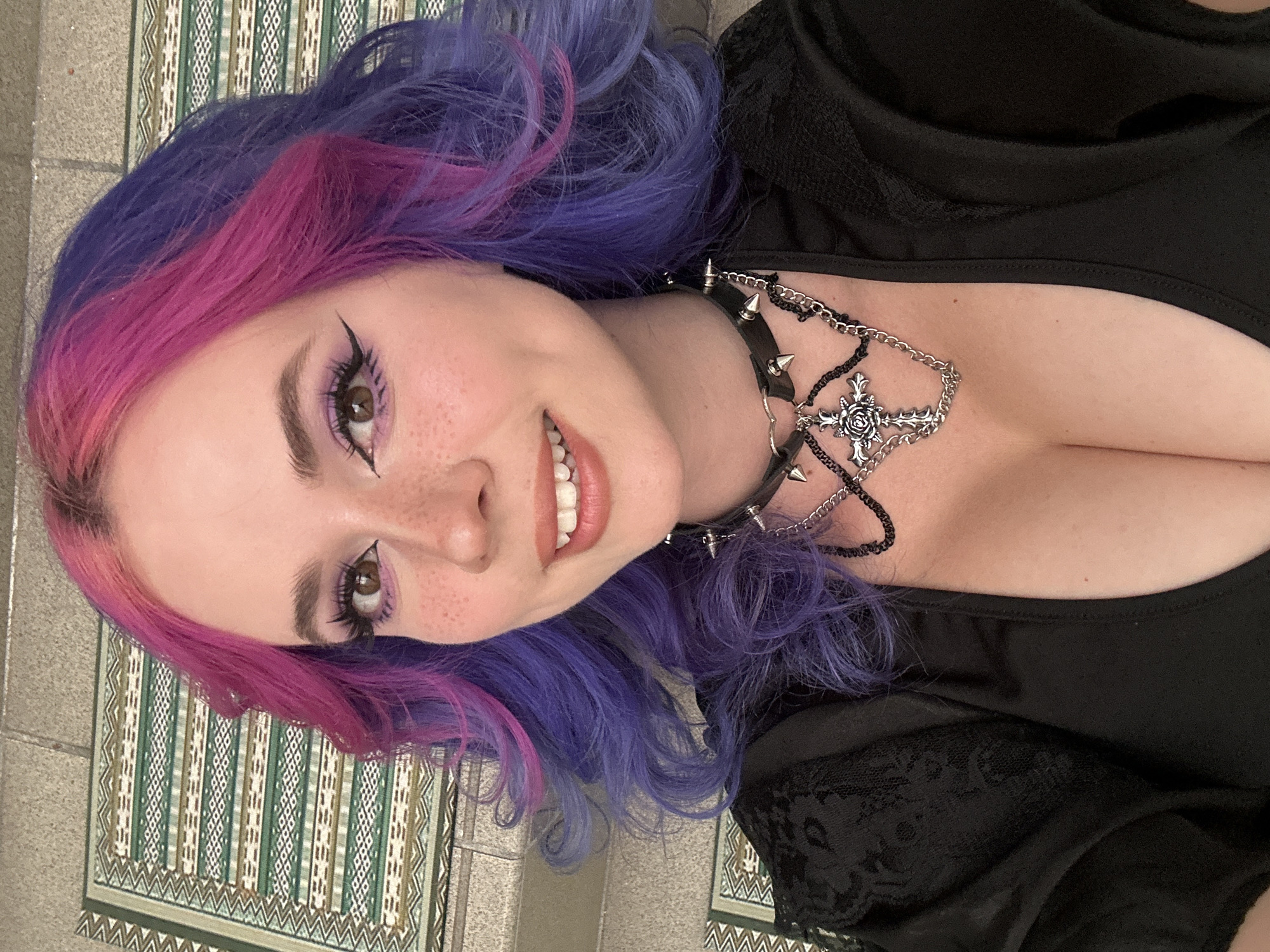 A young woman is depicted in a close-up, rotated portrait, smiling and looking directly at the camera. She has striking bi-colored hair, featuring vibrant fuchsia pink strands at the front and a deep purple color throughout the rest of her wavy hair. Her eyes are dark brown, enhanced with elaborate black winged eyeliner and purple eyeshadow, complemented by long, dark eyelashes. Freckles are visible across her nose and cheeks, and her lips are slightly parted in a wide smile, revealing white teeth.
She is wearing a black top, possibly a dress or blouse, with a subtle lace pattern visible on the shoulder/sleeve area to her right. Her neck is adorned with two distinct accessories: a black studded choker with silver spikes, and a layered silver chain necklace. This second necklace features a central pendant shaped like a cross, intricately detailed with a rose design, and includes black beaded sections.
The background consists of decorative tiles, possibly on a wall or floor, with a repeated geometric pattern in shades of green, white, and brown, arranged in vertical stripes. This setting suggests an indoor or semi-outdoor location, characteristic of architectural elements that might be found in the City of Zagreb, Croatia. The overall mood conveyed by her expression is cheerful and engaging. There is no visible text in the image.