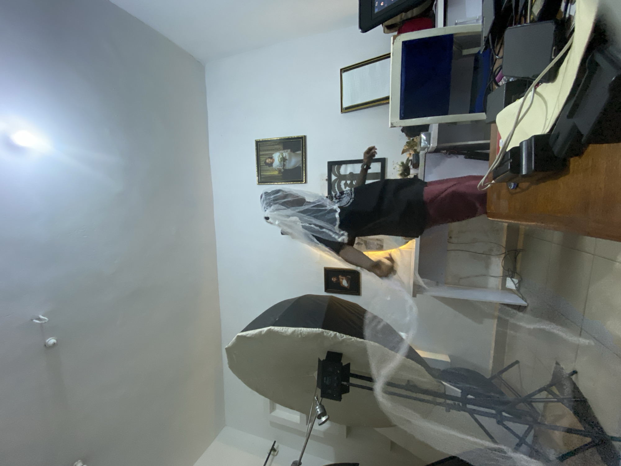 The image depicts an indoor scene, possibly a studio or a room in Bukuru, Nigeria. A person, wearing a black t-shirt and a reddish skirt, stands in the center of the frame, covered with a white veil. They are positioned near a large, white photography umbrella with a black backing, which is mounted on a stand. To the left, there's another piece of photography equipment. The background features white walls adorned with framed pictures, one of which appears to be a portrait of a woman in a wedding dress. There's also a desk or table with various items, including electronics and framed objects. The lighting suggests an artificial light source, possibly a studio light. The overall impression is of a creative space being used for photography or a related activity.