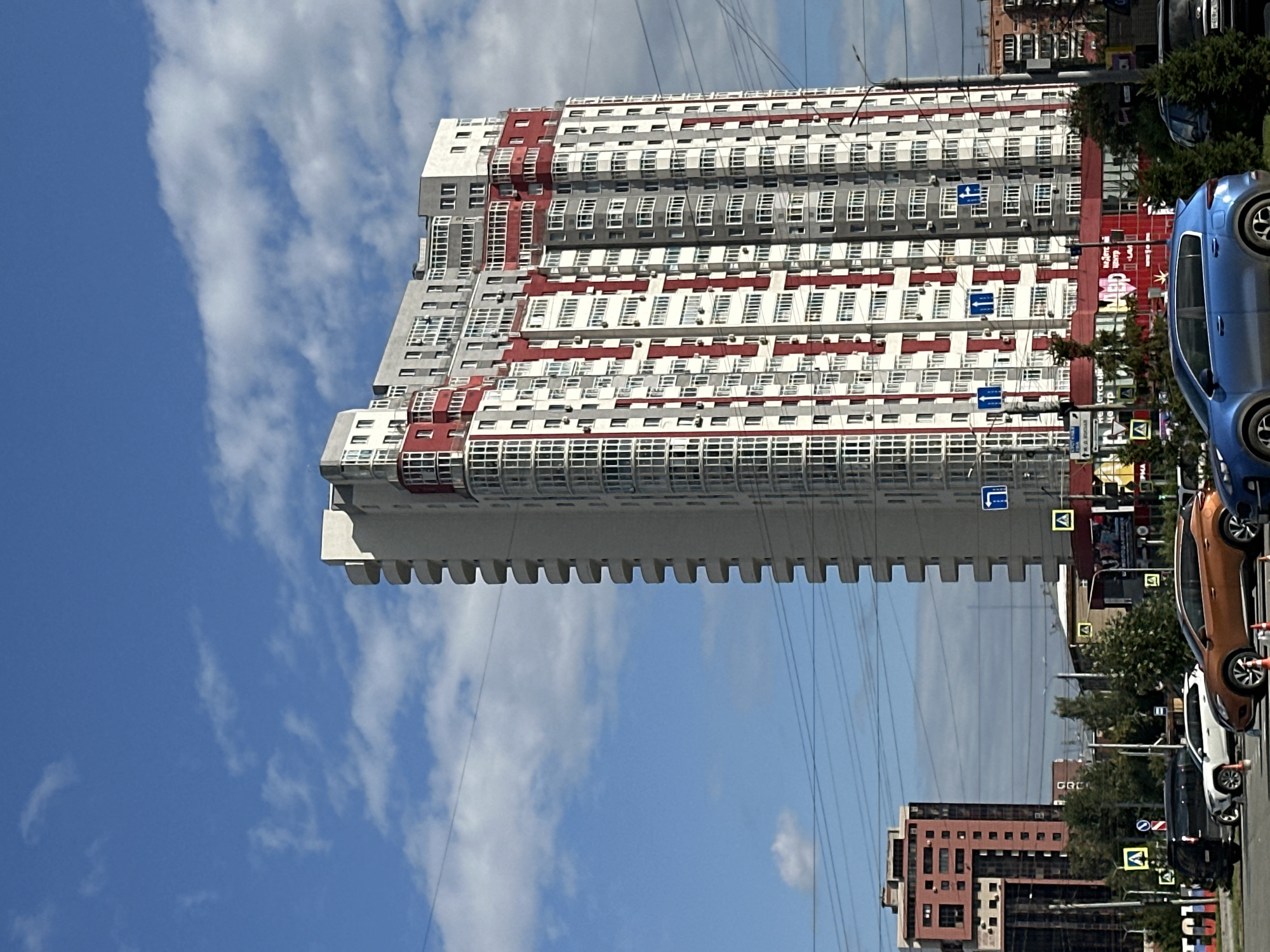 A tall, modern, residential building with a white and red facade dominates the center of the frame, set against a bright blue sky with scattered white clouds. The building has numerous balconies and many windows. To the left of this main building, a shorter, reddish-brown building with a distinct geometric shape is visible. In the foreground, a street scene unfolds with several cars parked and in motion, including a black SUV, a white SUV, an orange SUV, and a blue hatchback. Traffic lights and street signs are present, as are trees lining the road. The overall impression is a snapshot of an urban environment on a clear day.