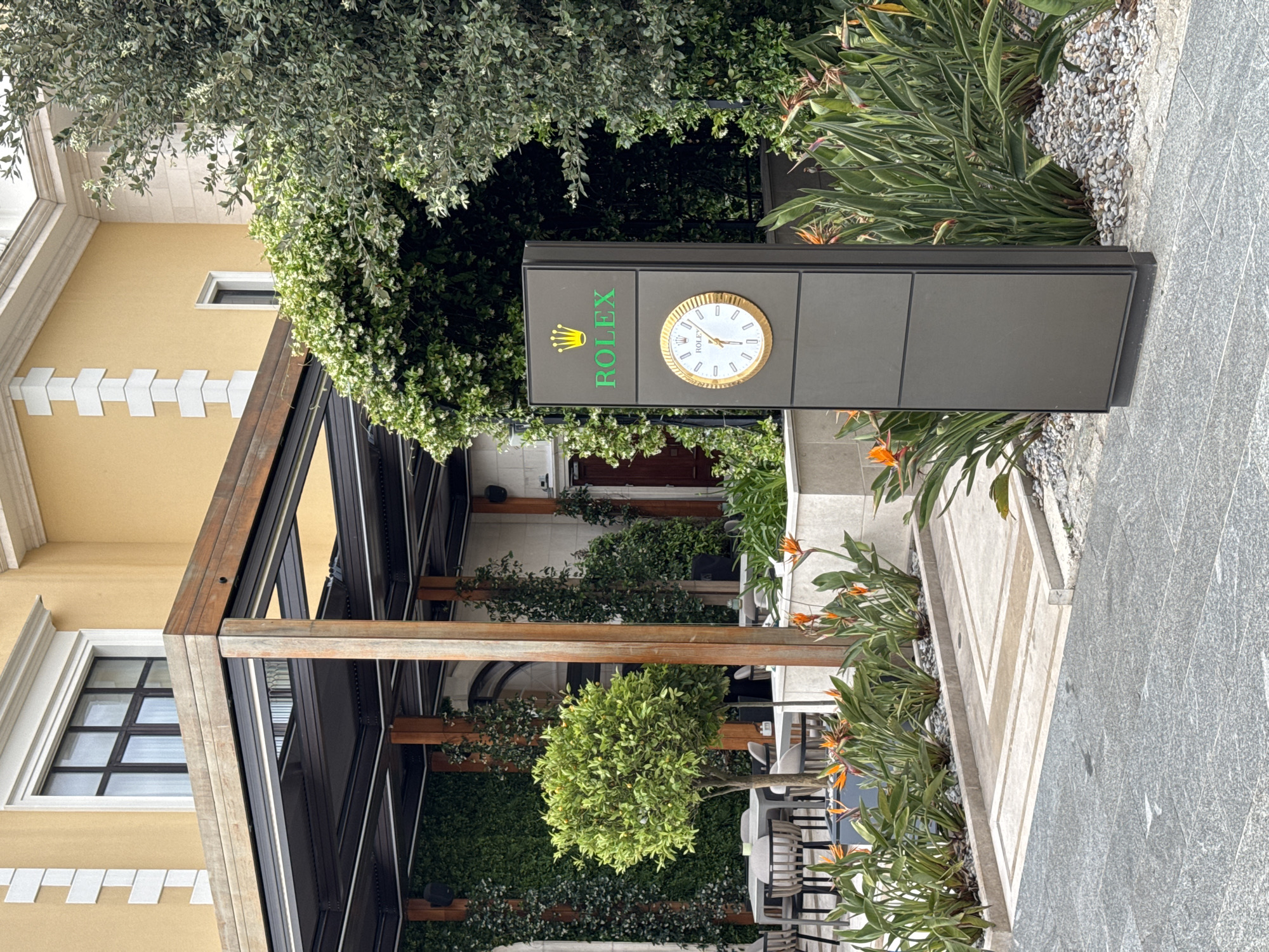 The image shows a Rolex clock sign in Tivat, Montenegro. The clock is part of a freestanding gray rectangular sign with the Rolex logo in green. The sign is surrounded by lush greenery and flowers, with a modern building visible in the background. The building has a pergola structure and black-framed windows. Tables and chairs suggest a restaurant or cafe setting. The ground is paved with light gray stone.