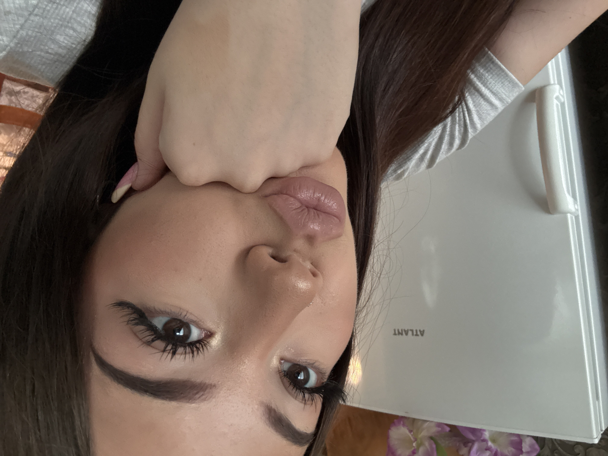 A close-up, angled view of a person's face, likely female. The person's hand is positioned against their face, distorting their lips. The person has dark hair, defined eyebrows, and appears to be wearing eye makeup. A white object with the word "ATLANT" is visible in the background. The exact location of the scene is not specified in the image itself. There is also flowers in the background, out of focus.