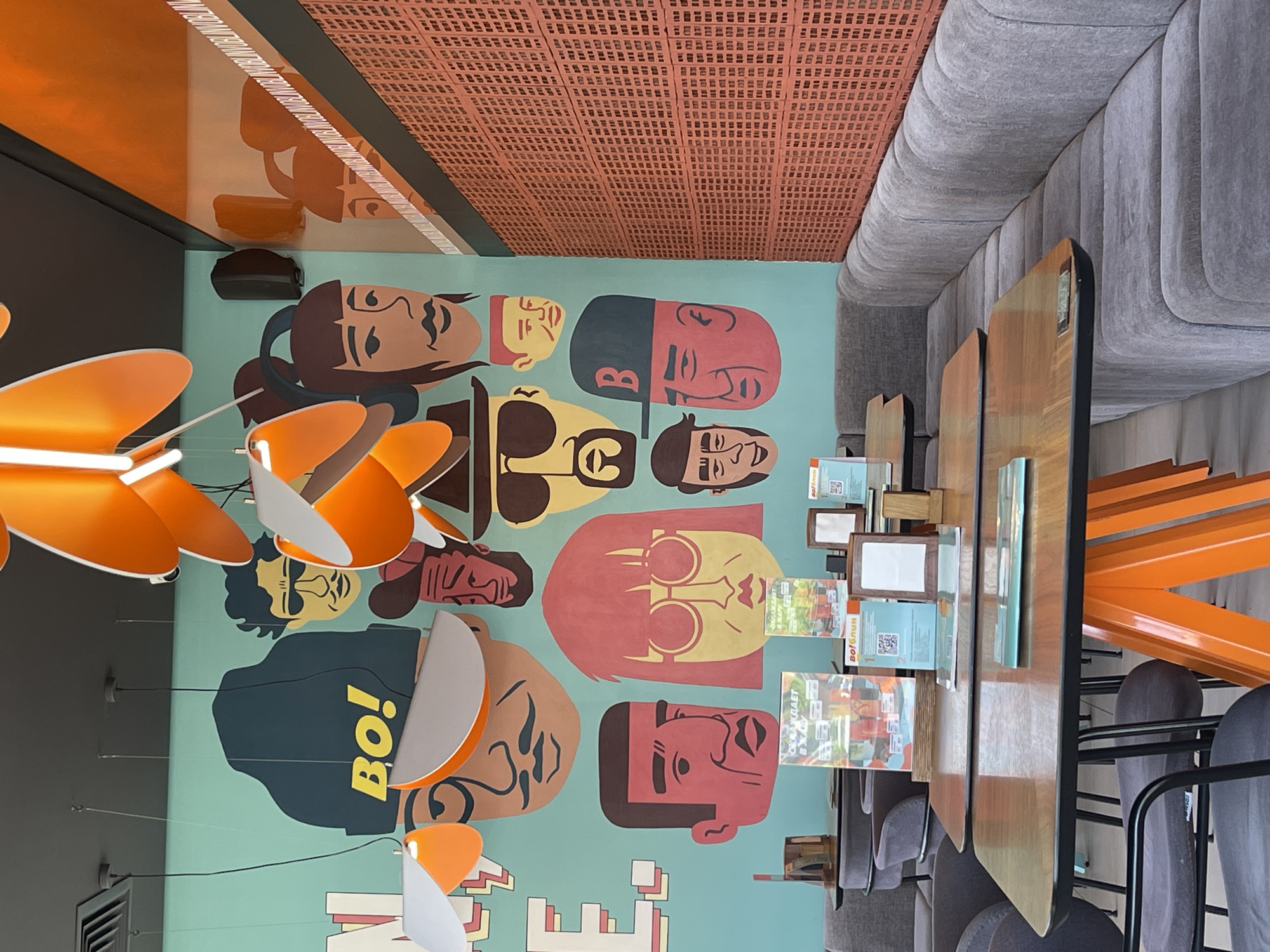 The image depicts an interior shot of a commercial establishment, likely a restaurant or cafe, located in Stavropol, Russia. A mural adorns a wall, featuring a series of stylized, colorful portraits of individuals. Above the mural, the ceiling has a distinct pattern, possibly constructed of terracotta blocks.
The foreground contains a row of tables with menus or informational cards on display. Chairs are neatly arranged alongside the tables. Overhead, a modern, orange and white light fixture hangs. To the right of the image, part of a gray upholstered banquette is visible.