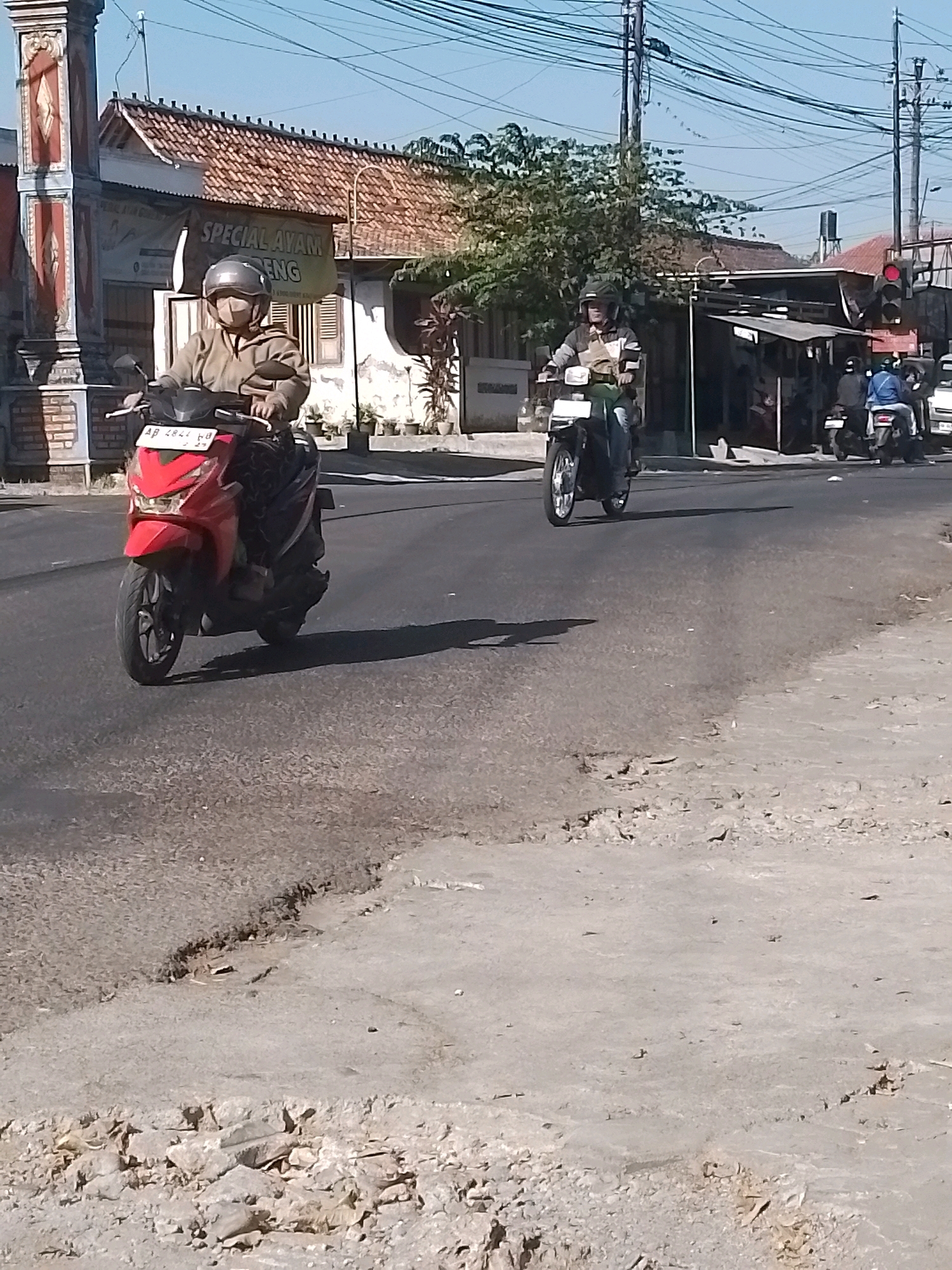 On a sunny day in Wonosari, Indonesia, two individuals are seen riding motorcycles on a road. The first motorcycle, a red scooter, is closer to the foreground. The rider is wearing a light brown jacket, a helmet, and a face mask. The motorcycle has a license plate that reads "AB 2821 HB". The second motorcycle, a dark-colored scooter, is further down the road. The rider is wearing a helmet and a jacket with striped sleeves. Behind the motorcycles, buildings with red tile roofs are visible, along with trees and overhead power lines. A banner that reads "SPECIAL AYAM GORENG" is hanging on one of the buildings. To the right, a traffic light is red, and other motorcycles are present. The road surface shows signs of wear and tear, with patches of dirt and gravel.
