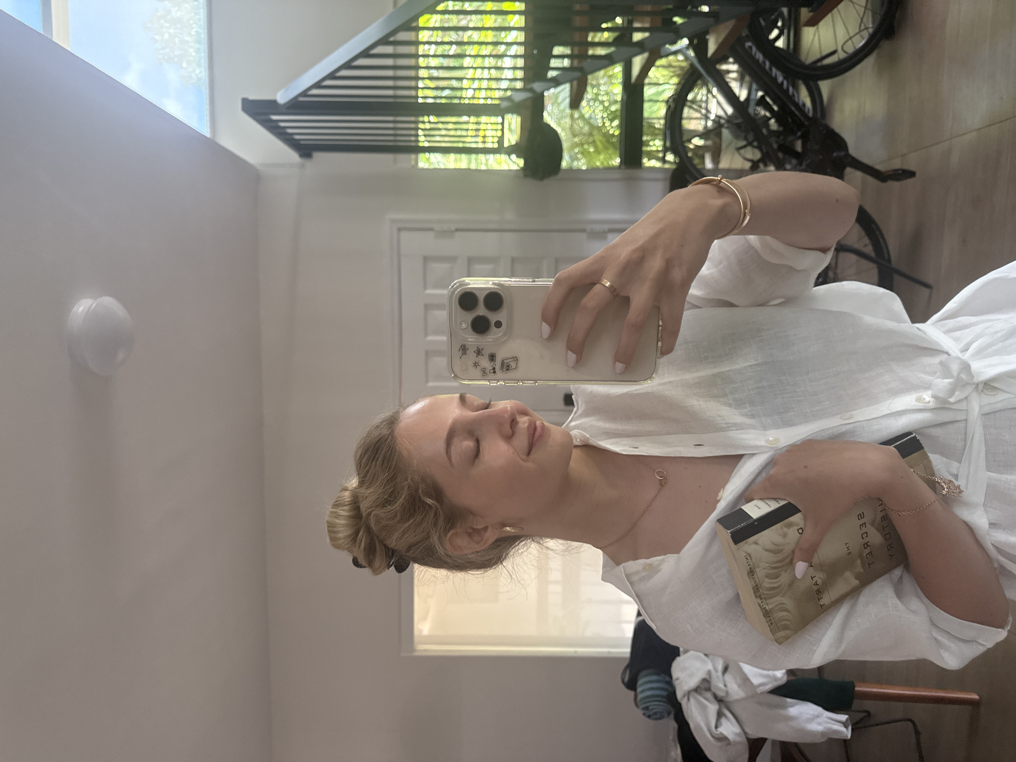 A young woman with blonde hair in a bun is taking a mirror selfie in a well-lit, modern home. She is wearing a white linen shirt and holding a book titled "The Secret History" by Donna Tartt. The background shows a contemporary staircase with black railings, bicycles, and a large window with greenery visible outside. The overall ambiance suggests a relaxed and stylish interior. The image does not provide any specific clues about its location within the city of Miami, United States of America.