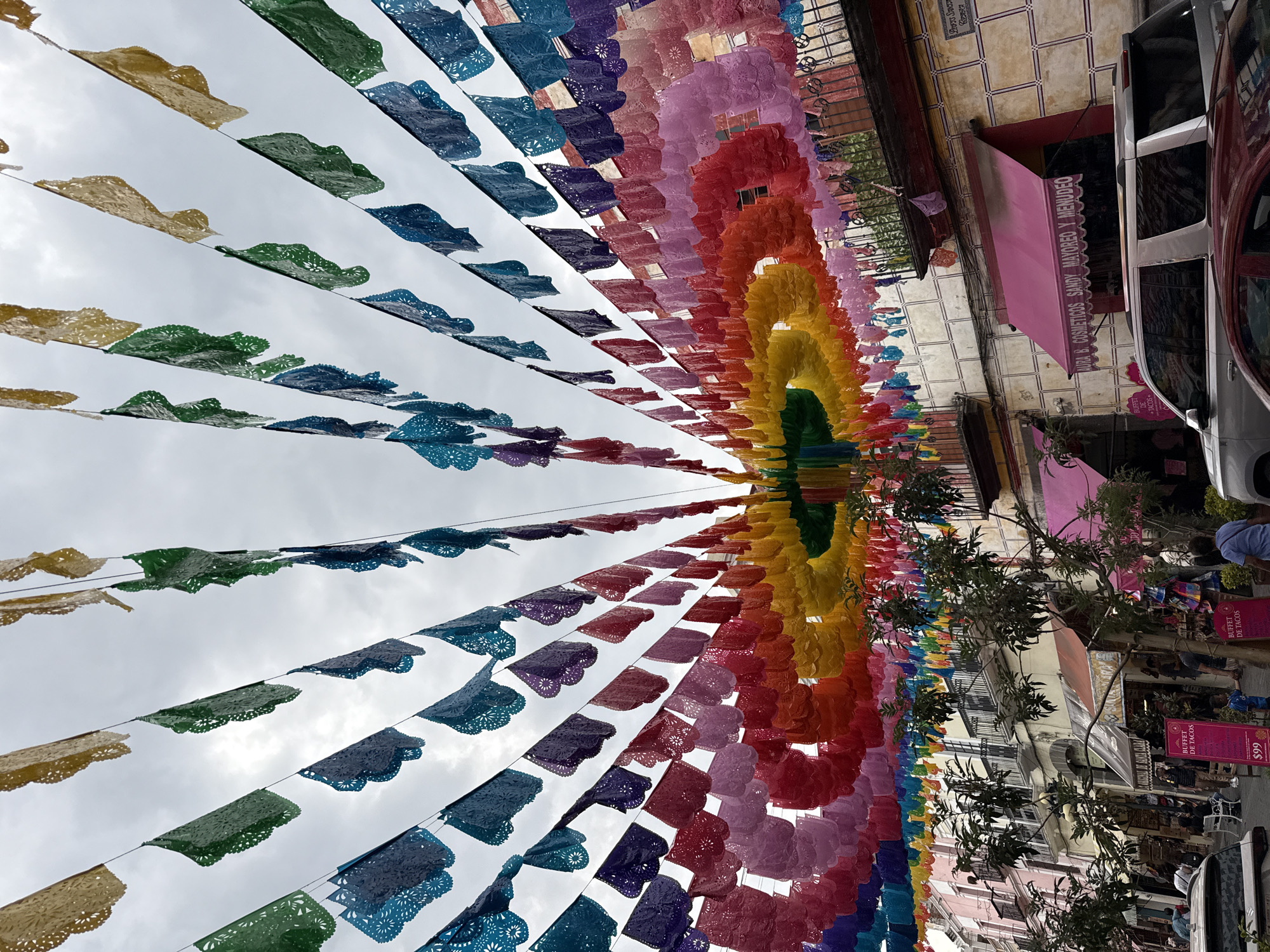 This vibrant image captures a street in Puebla, Mexico, adorned with countless colorful *papel picado* banners strung overhead. The perspective looks upward along the street, creating a tunnel-like effect with the intricate paper decorations, which transition through a rainbow spectrum of green, yellow, orange, red, pink, purple, and blue. The sky above is cloudy white/grey, providing an even, soft light.
On the right side of the image, traditional buildings line the street, featuring multi-story facades, balconies with wrought iron railings, and tiled walls. Storefronts are visible below, some with bright pink awnings. A silver vehicle, possibly a van or SUV, is partially parked along the street. Several small trees with green foliage are interspersed among the buildings, and a few people can be seen walking in the distance, though their details are indistinct.
The numerous *papel picado* suggest a festive atmosphere, common in Mexican celebrations. Visible text includes "SANDY MAYOREO Y MENUDEO" (wholesale and retail) for "COSMETICOS" (cosmetics) on a pink awning. Red signs clearly advertise "BUFFET DE TACOS" for "$99" (likely Mexican pesos). Another partial text "LOFT'S SANT" can also be seen.