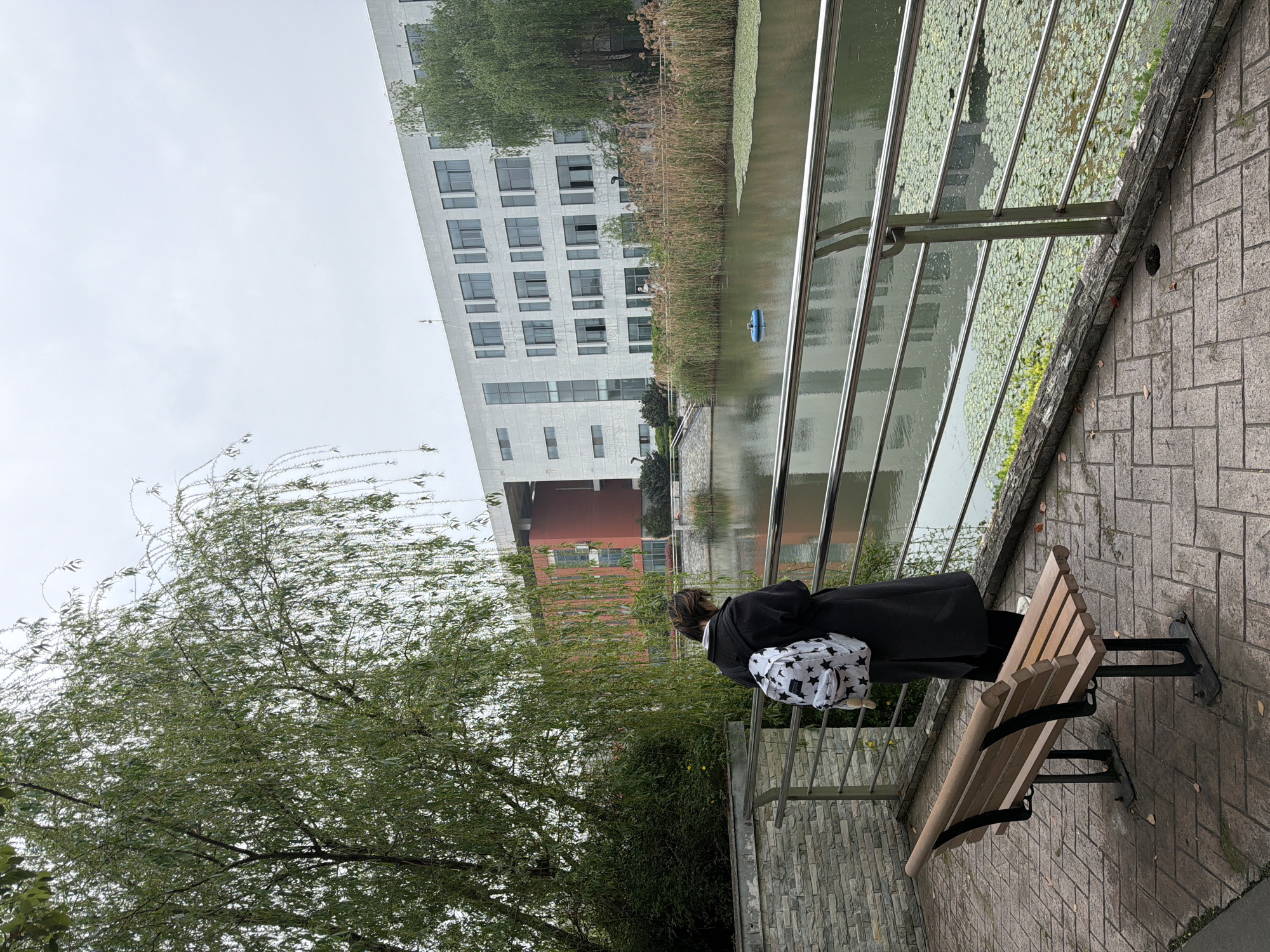 A medium shot captures a person standing behind a park bench along a river or canal in Jiangning District, China. The person, wearing a black coat and carrying a white backpack with black stars, stands near a metal railing bordering the water. Across the water is a large, modern building with a white facade, visible windows, and some red or brown elements. Reeds and lily pads grow along the water's edge. A tree with drooping branches is on the left, partially obscuring the view of a lower building. The bench is wooden with black metal supports, and the ground is paved with brick. The sky appears overcast.
