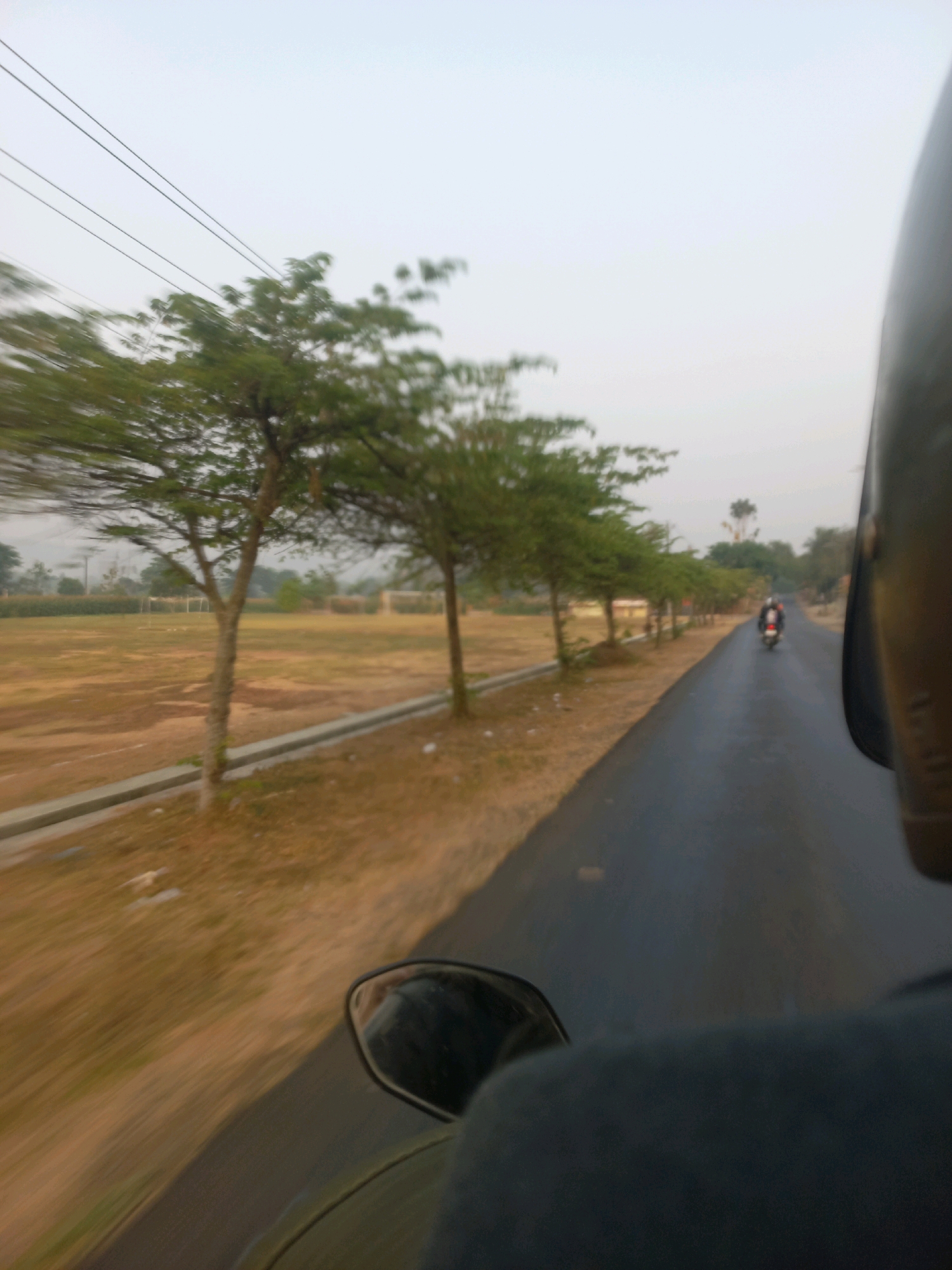 A first-person perspective shot taken from a moving motorcycle on a paved road in Tanjungsari, Indonesia. The shot captures a line of trees on the left side of the road, with a dry, brown field behind them. Power lines are visible in the upper left corner of the frame. In the distance, two motorcycles are traveling in the same direction. The image is blurry due to motion, with the helmet of the rider in the foreground obscuring a portion of the right side of the frame.