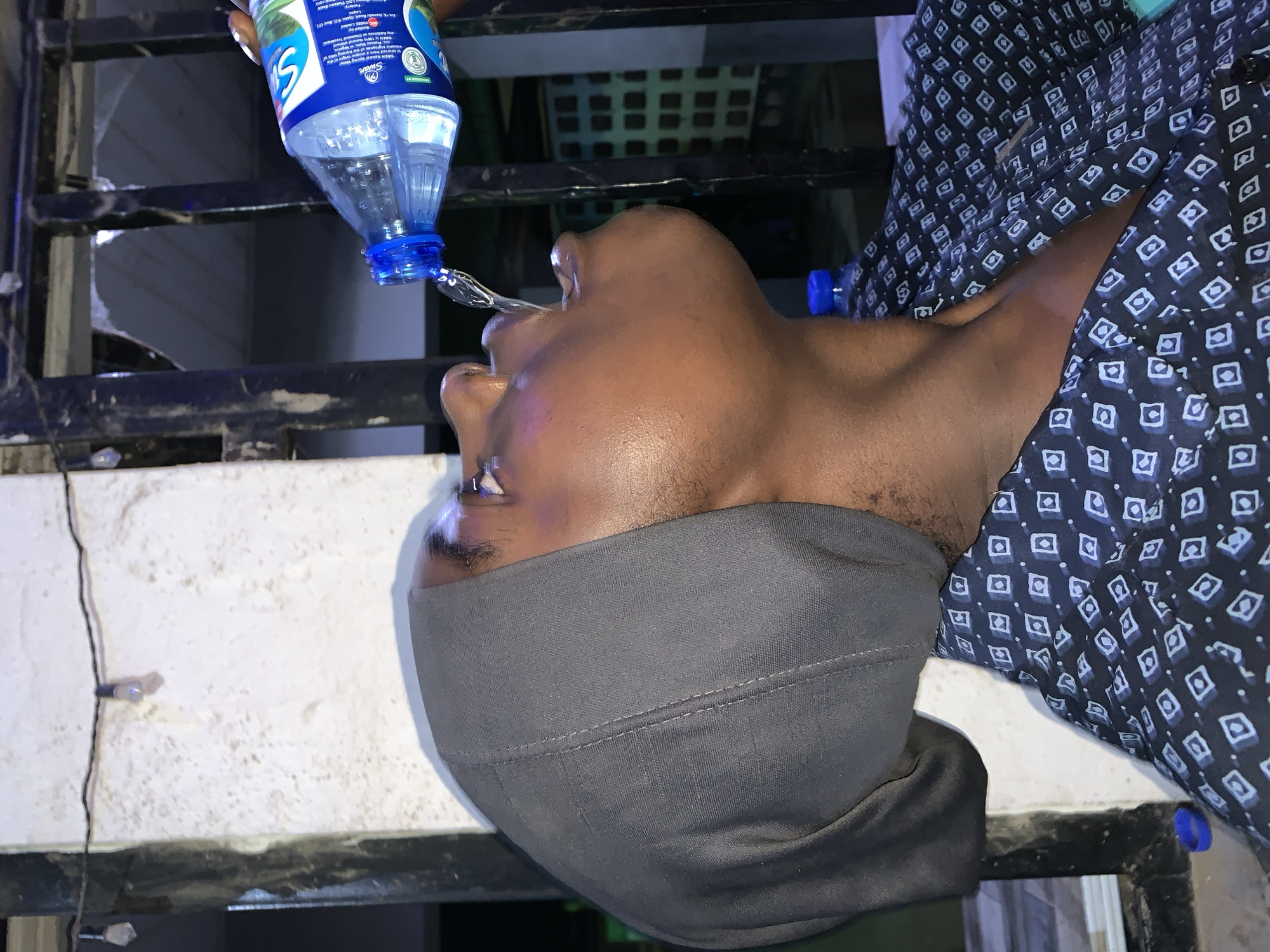 A dark-skinned person wearing a gray cap and a patterned button-down shirt is drinking water from a plastic bottle. The person's head is tilted back to allow the water to pour directly into their mouth. The scene takes place near a barred window or railing. No location data is available in the provided information, thus the image cannot be geographically located within New Stadium, Nigeria.