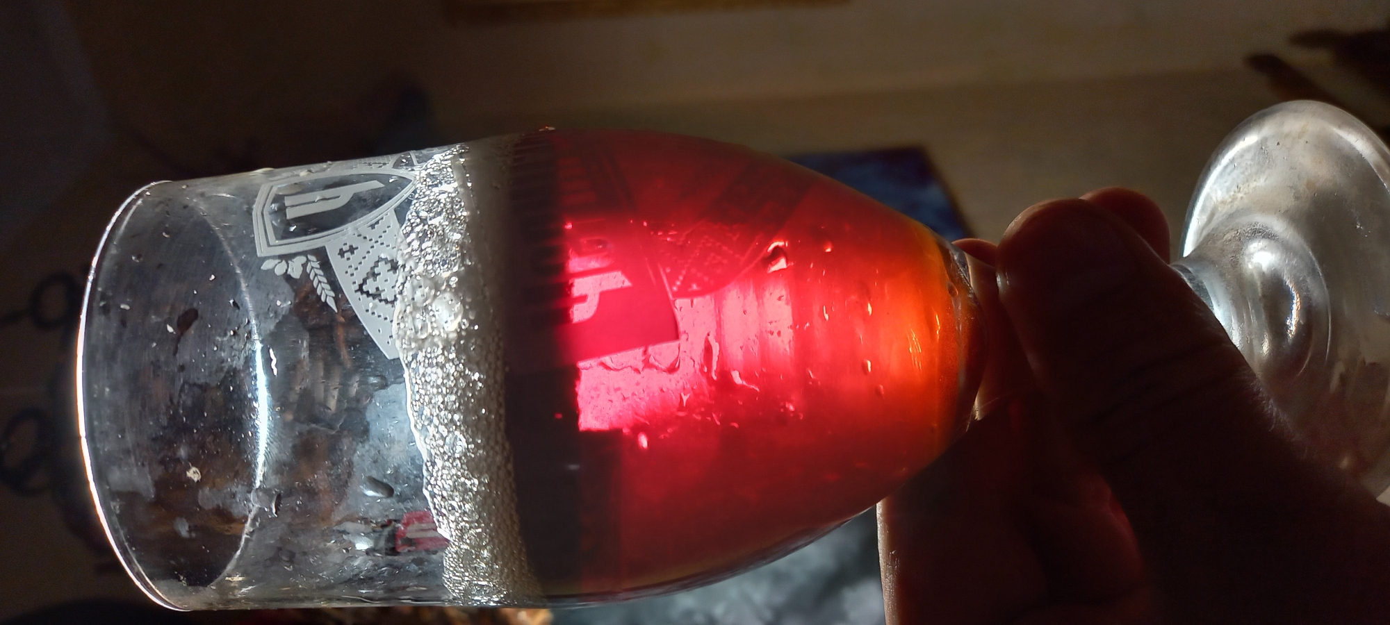 A stemmed glass, approximately two-thirds full of a translucent reddish-orange liquid, is held by a human hand. The liquid has a prominent layer of off-white foam on its surface. Condensation and numerous water droplets adhere to the exterior of the glass. An etched or printed emblem featuring a shield with a stylized letter 'Ψ' (Psi) or similar symbol, a sprig of leaves or wheat, and a patterned base, is visible on the upper half of the glass. The liquid glows intensely due to strong backlighting, which also silhouettes the hand holding the glass. Dark, vertical reflections are discernible within the red liquid. The lower portion of the liquid appears more orange, possibly due to light refraction or sediment. The background is dark, blurred, and indistinct, indicating an indoor setting. The specific location within the city of Kryvyi Rih, Ukraine, cannot be determined from the visual evidence.