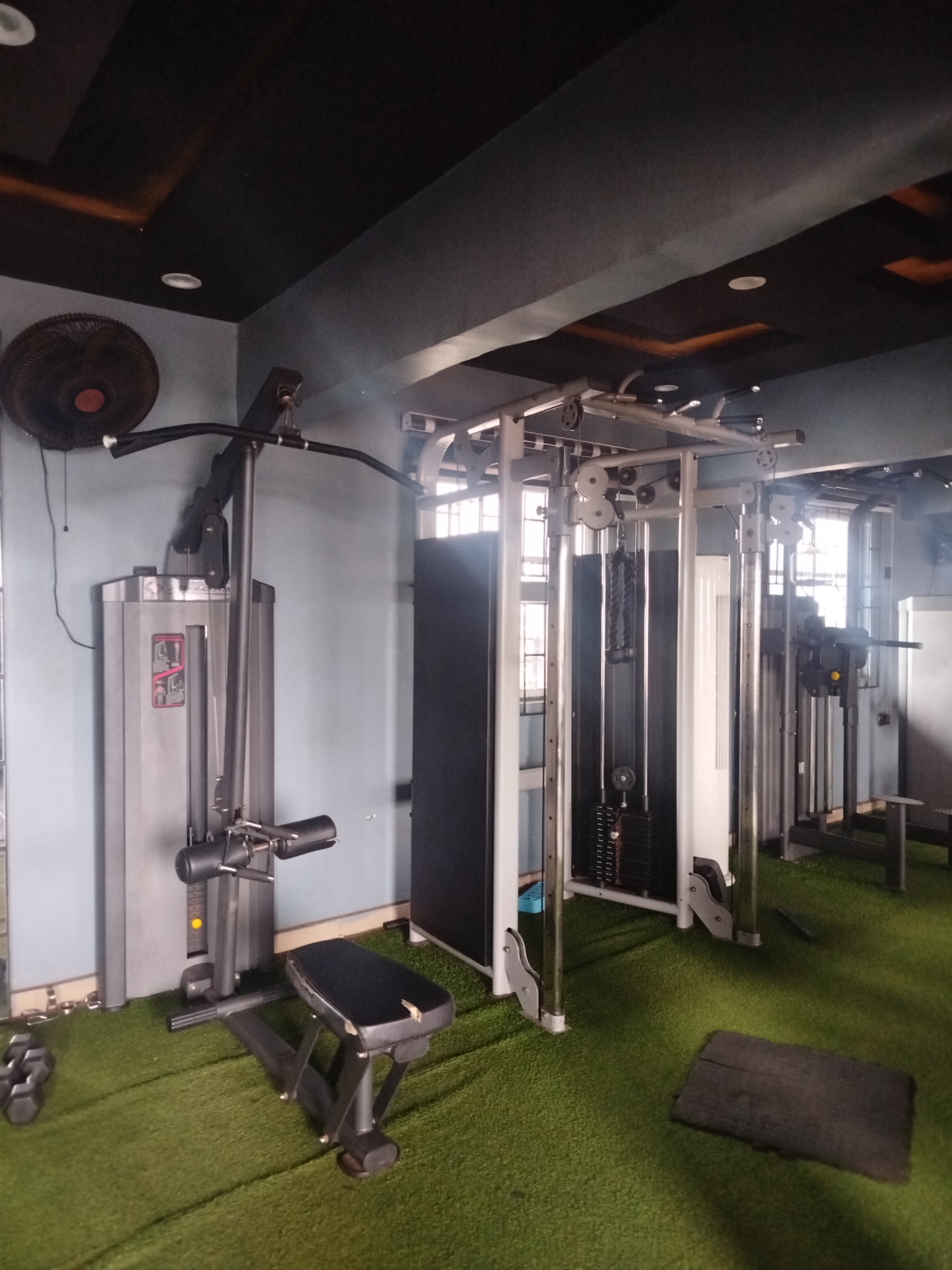 The image shows the interior of a gym in Uselu, Nigeria. The gym features artificial turf flooring and is equipped with various exercise machines, including a lat pulldown machine, a cable crossover machine, and weight racks. A wall-mounted fan is visible in the upper left corner. The lighting appears to be a mix of natural light from windows and artificial lighting from the ceiling. There are no people in the image.