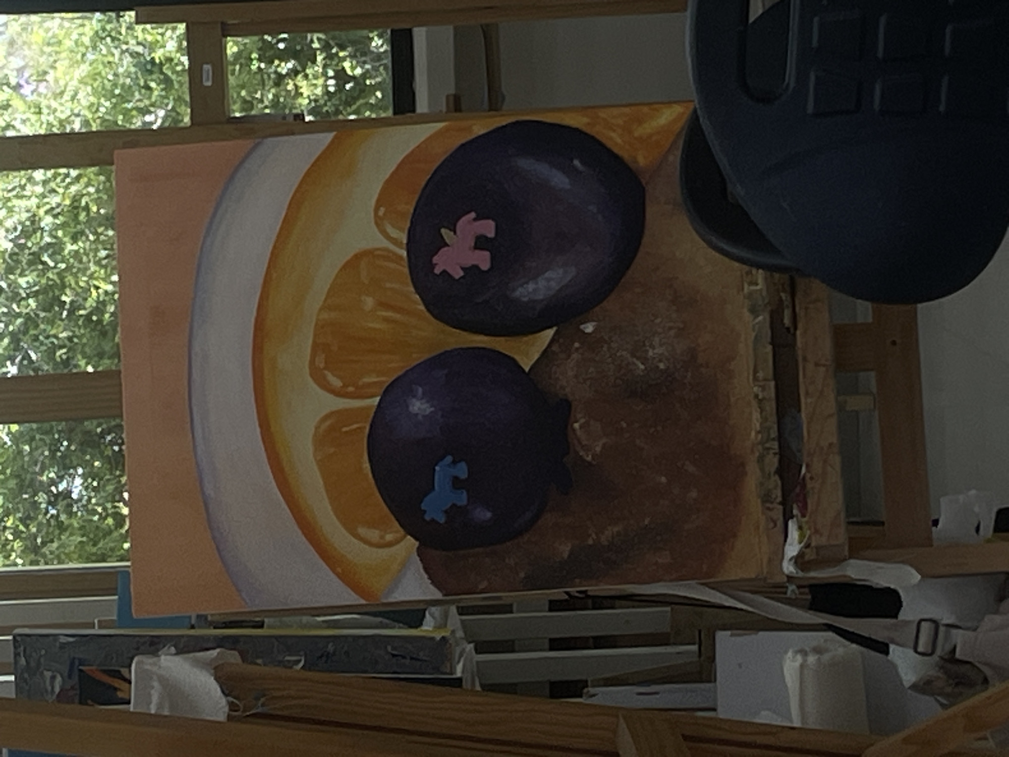 The media file captures a close-up view of an artwork in what appears to be an art studio, possibly in San Andrés Cholula, Mexico. The central subject is a vibrant painting featuring a still life arrangement. The painting depicts several slices of citrus fruit, likely oranges or lemons, rendered in bright yellow and orange tones with visible segment lines. A white or light grey rim of a plate or bowl is partially visible on the left side, framing the citrus. Two large, dark purple fruits, resembling blueberries or mangosteens, are prominently placed on top of the citrus slices. Each of these purple fruits has a distinct, lighter-colored marking on its surface; one has a pink, almost jigsaw-piece-like shape, and the other has a similar shape in light blue. The background of the painted composition is a textured brown.
The painting is mounted on a wooden easel, positioned in front of a window. Through the window, lush green foliage of trees or bushes is visible, suggesting an outdoor natural environment and indicating a daytime setting with ample natural light. Below the painting, various objects associated with an art studio are visible, including what look like other canvases or boards, some rolls of white material (possibly paper towels or rags), and other indistinct art supplies. A dark, irregularly shaped object, possibly part of a camera or equipment, is partially visible in the upper right corner of the frame. No discernible text is visible in the image. The scene conveys a quiet, creative atmosphere within an artist's workspace.