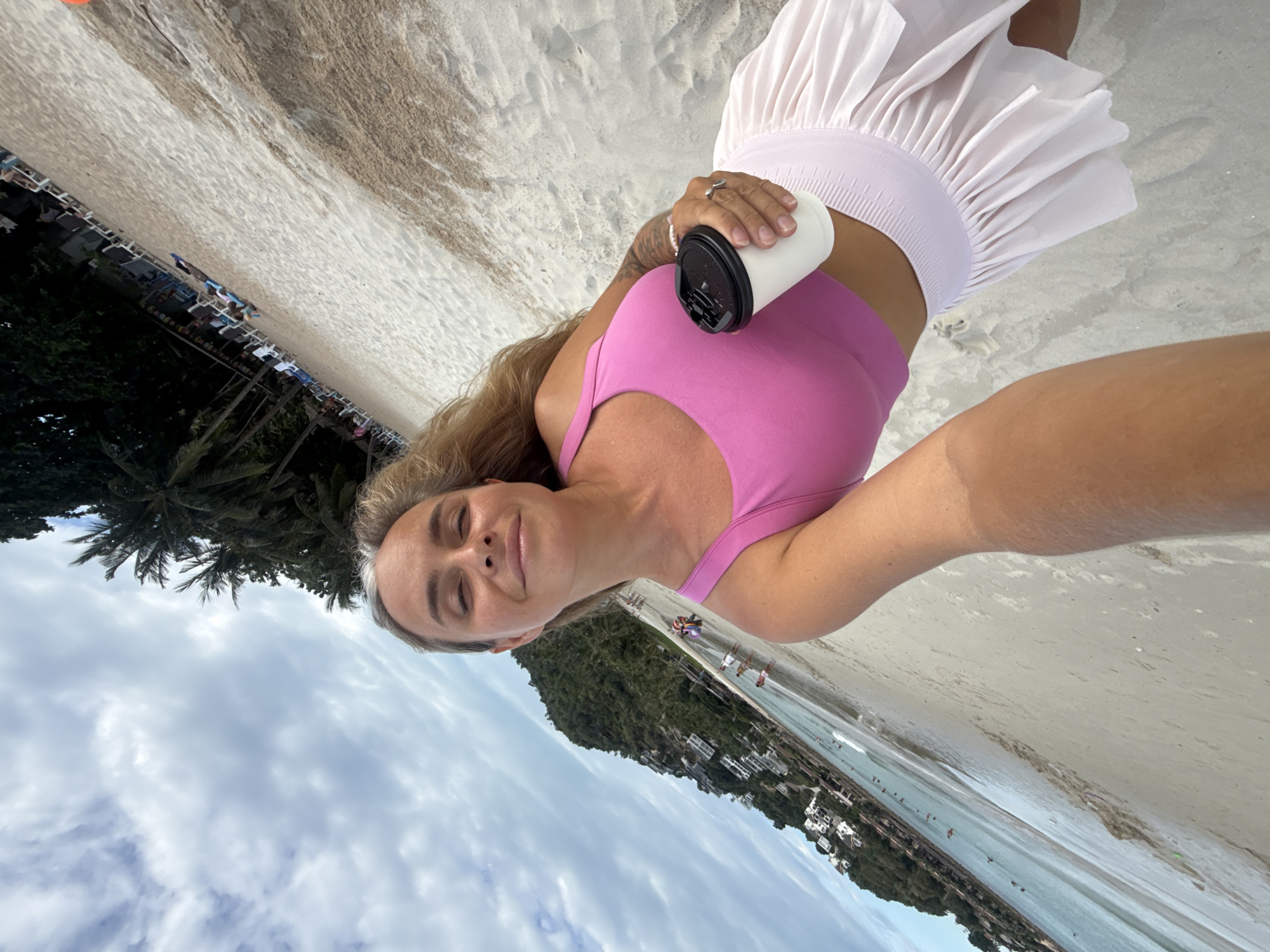 A woman is taking a selfie on a beach in Ko Samui District, Thailand. She is holding a white cup with a black lid in her right hand and wearing a pink tank top and a light pink pleated skirt. Her eyes are closed and she is smiling. The beach is sandy and there are palm trees in the background. There are buildings on a hill to the left of the image, and people are visible in the water and on the beach. The sky is cloudy.