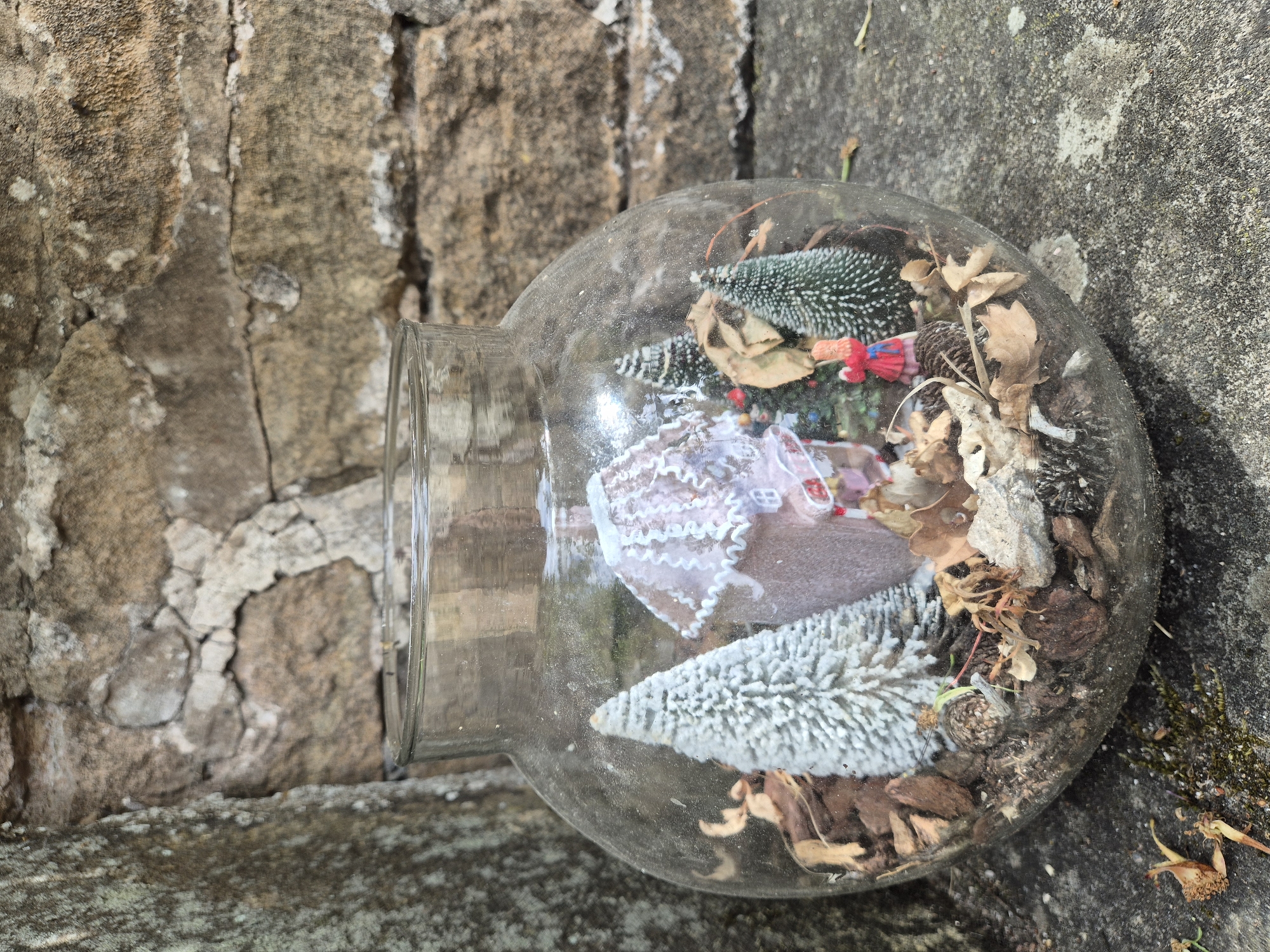 A glass terrarium containing miniature Christmas-themed decorations sits on a textured concrete surface next to a rough stone wall. The terrarium, which is spherical in shape with a wide opening, contains a small pink gingerbread-style house adorned with white icing, two bottle-brush trees with a frosted appearance, a small red-dressed figurine, and various natural elements such as pinecones, bark, and dried leaves. The background features a wall constructed of irregular, weathered stones. The lighting suggests an outdoor setting, possibly in a garden or courtyard. The image does not depict any specific location within the city of Gedling, United Kingdom.