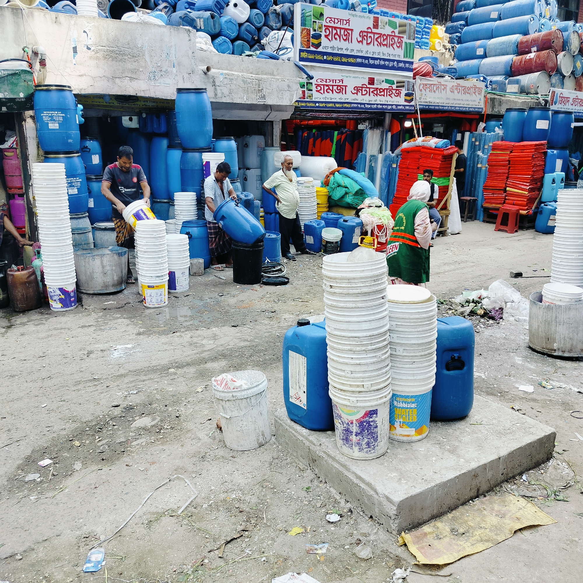 This image depicts a commercial area in Dhaka, Bangladesh, specifically in the Kitchen Market area of Kawran Bazar. The environment is cluttered and appears to be a wholesale or retail outlet for industrial containers, barrels, and plastic buckets.
Several individuals are present: two men are working with large blue plastic barrels, seemingly transferring or manipulating them. An older man stands nearby, observing, while another person, possibly a woman, wearing a green vest with the word "LEEN" printed on it, stands with their back to the camera, observing the scene.
Stacks of white plastic buckets dominate the foreground and the right side of the image. Additionally, large blue plastic barrels are stacked high in the background, and some are arranged around the people in the middle ground. Red tarp sheets are visible in the right middle-ground.
The ground is uneven, dusty, and littered with debris, suggesting a busy, possibly outdoor market setting. Banners advertising businesses like "Hamza Enterprise" and other names written in Bengali are visible above the storefronts, indicating the commercial nature of the area.