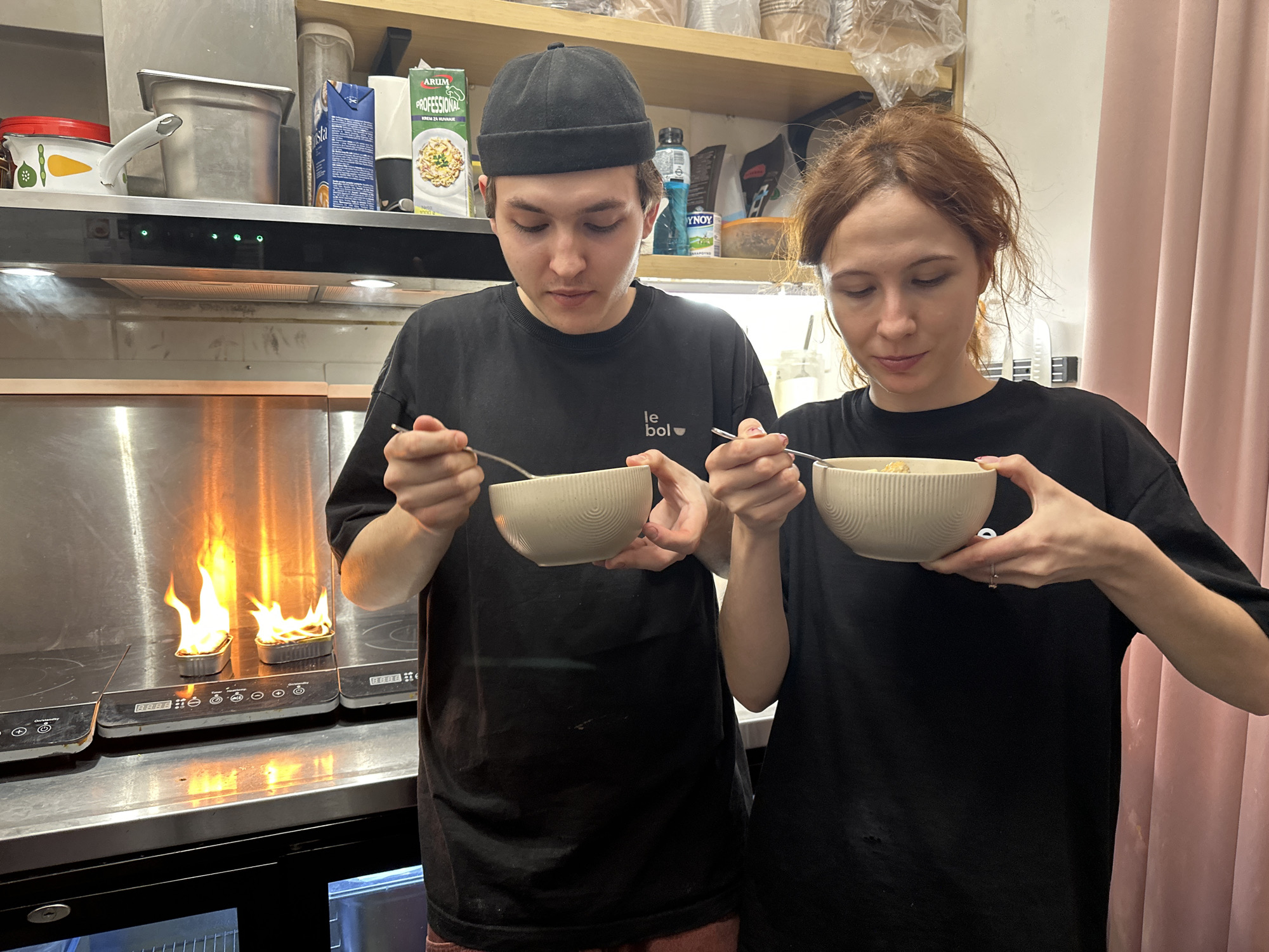 Two people, a man and a woman, stand side-by-side, each holding a bowl and a spoon. Both are wearing black t-shirts. The man is on the left, wearing a black cap, and the woman is on the right, with reddish-brown hair. They are looking down at their bowls, seemingly about to eat.
Behind them, a metal counter is visible, with two metal containers on a stovetop burner with flames coming out of them. Above the counter is a shelf with various items, including food containers and a bottle of water. A pink curtain hangs on the right side of the frame. The location is a kitchen, possibly in a restaurant.
While this cannot be confirmed with certainty based on visual evidence alone, the t-shirt worn by the man has "Le Bol" written on it, a known restaurant chain in Novi Sad, Serbia, suggesting that this photo was taken in that city.
