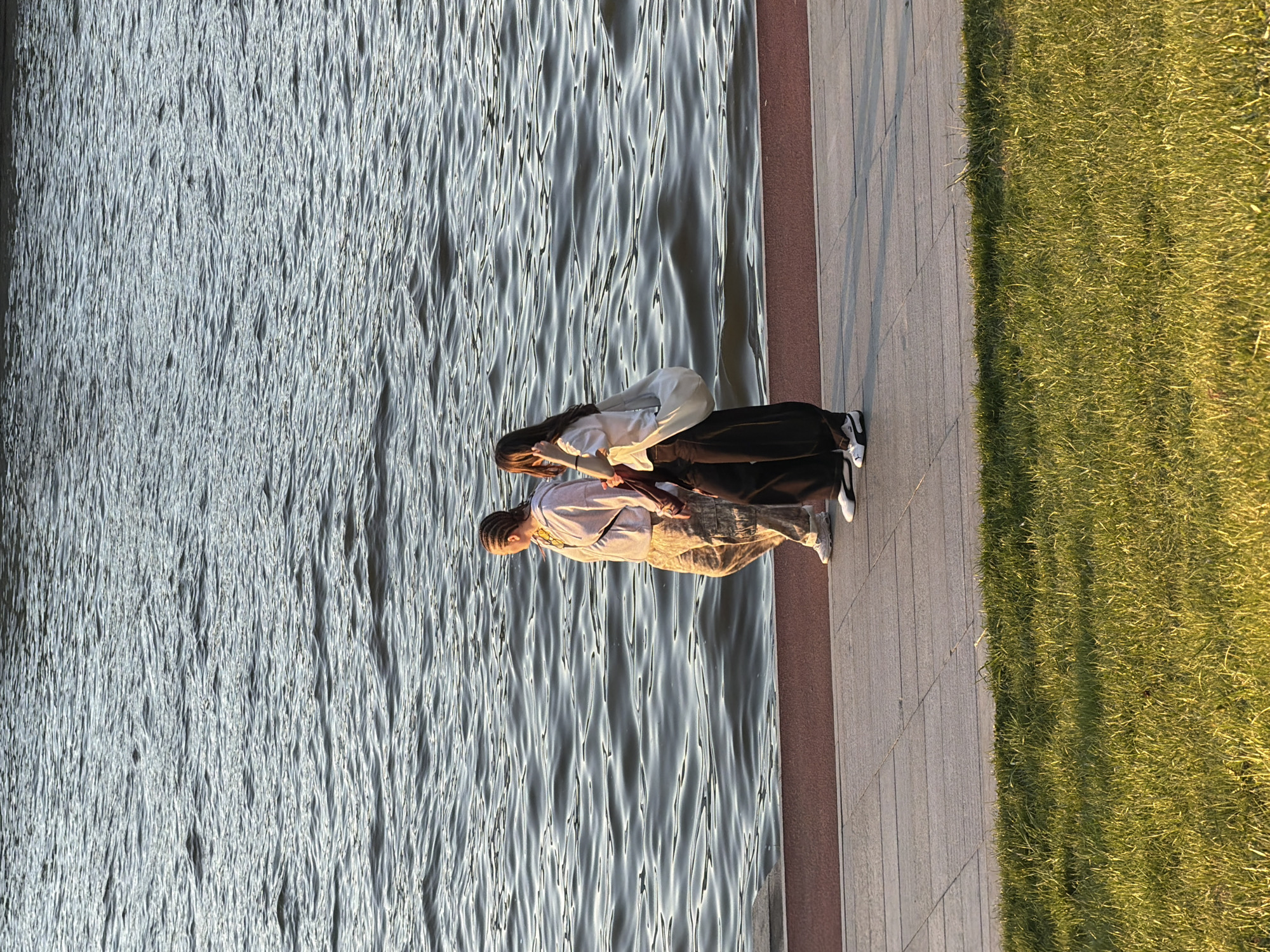 Two individuals are standing near the water's edge on a paved walkway. One individual wears a light-colored shirt and camouflage-patterned pants. The other individual wears a white shirt and black pants. The water is to their left, and grass is to their right. The location is most likely in Moscow, Russia.