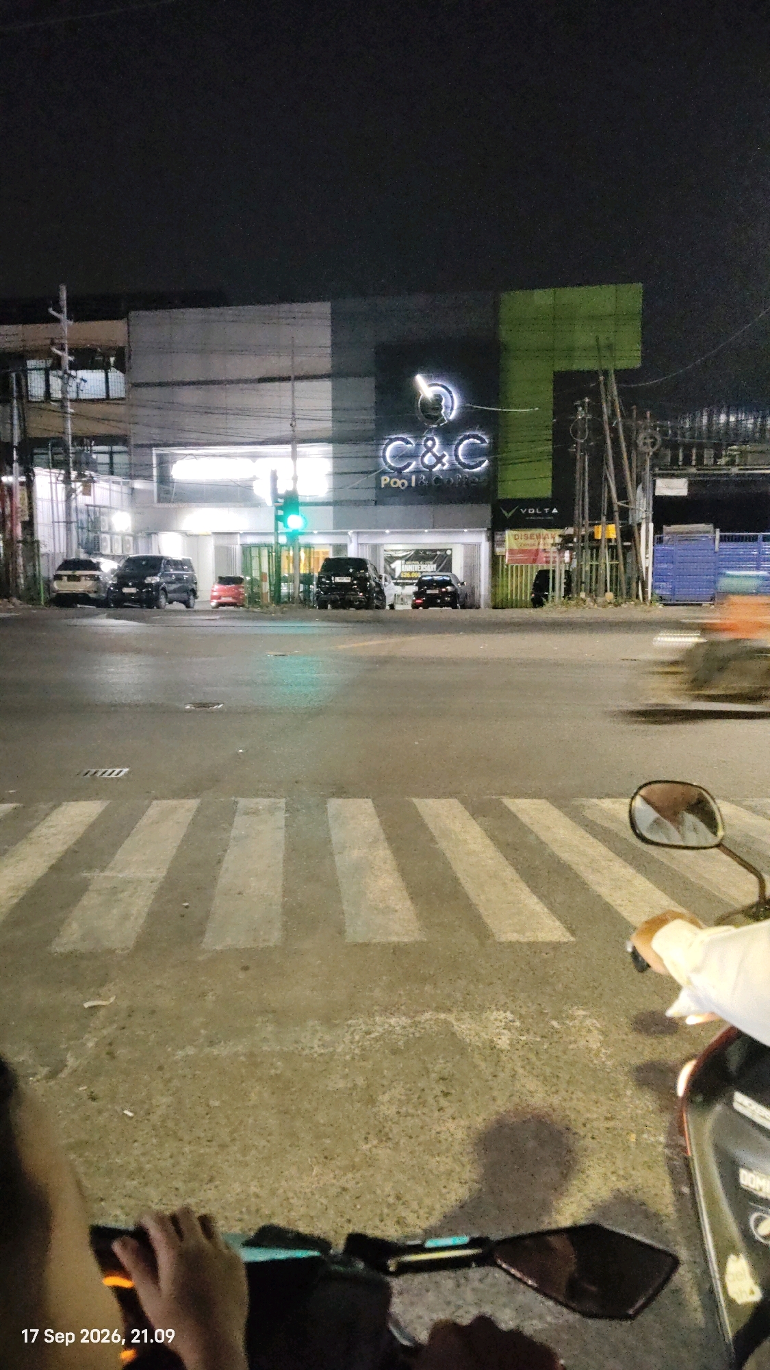 A night-time street scene in Surabaya, Indonesia, shows a busy intersection with several cars parked and driving. In the background, a building with illuminated signage for "C&C Pool & Coffee" is visible. The street is wet, likely from rain, and a pedestrian crossing is marked by white stripes. A motorcycle is in the foreground, with its rider's hands on the handlebars, suggesting they are either waiting at a traffic light or stopped. The date and time "17 Sep 2026, 21.09" are superimposed on the bottom left of the image. The overall atmosphere is urban and lively, typical of a commercial area at night.
