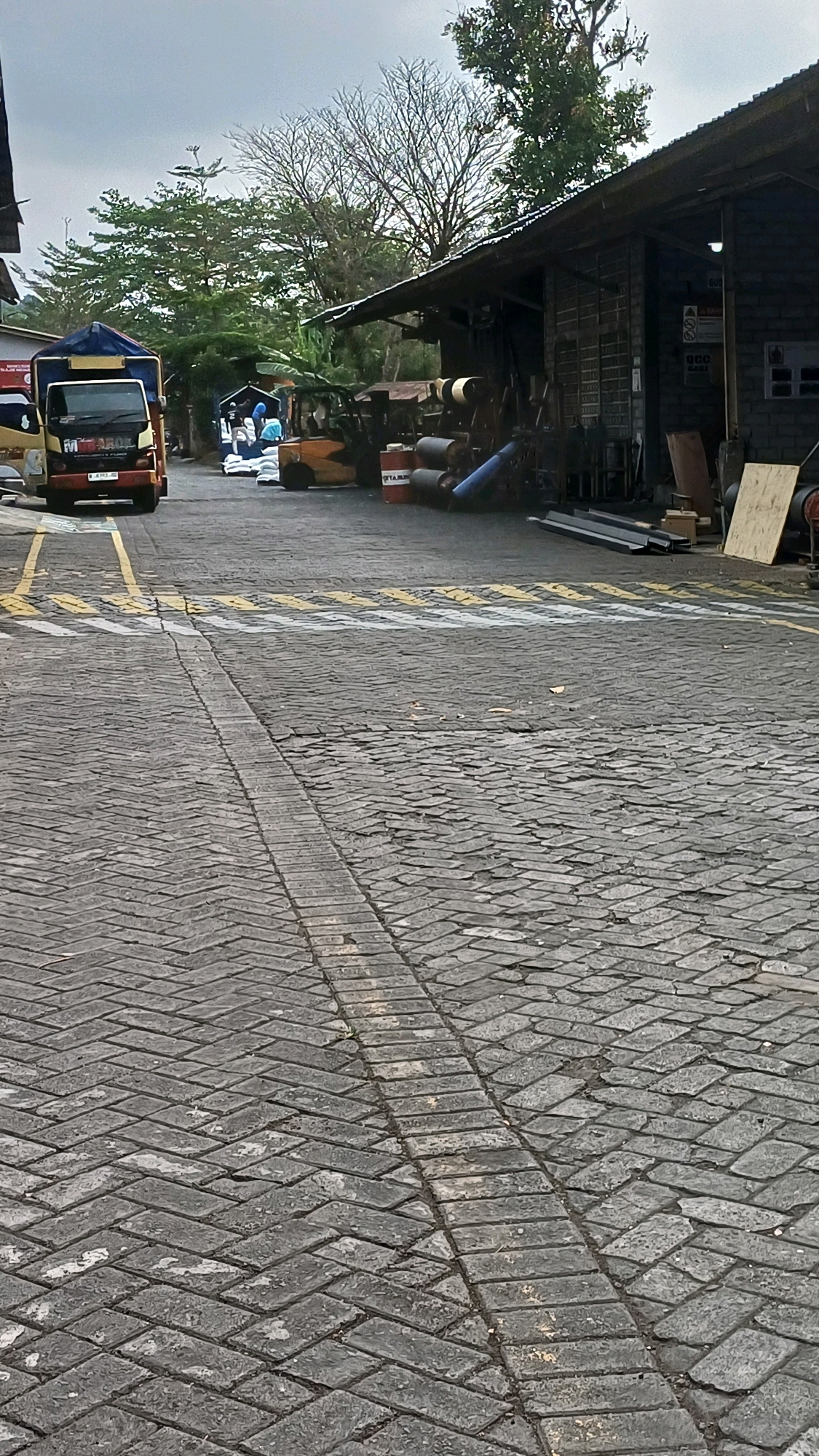 In the city of Kranggan, Indonesia, a paved industrial area is depicted. A truck with the logo "MITAROL" is parked on the left side of the image. Behind it, several individuals in blue and white attire are seen near a forklift, possibly involved in unloading or loading cargo. The ground is covered in a herringbone pattern of grey bricks. In the background, a long, low building with a corrugated metal roof stands, with various materials and equipment visible under its overhang. Trees with sparse foliage and a few green leaves are present, suggesting a dry season or that the trees are naturally bare. The sky is overcast, casting a diffused light over the scene.