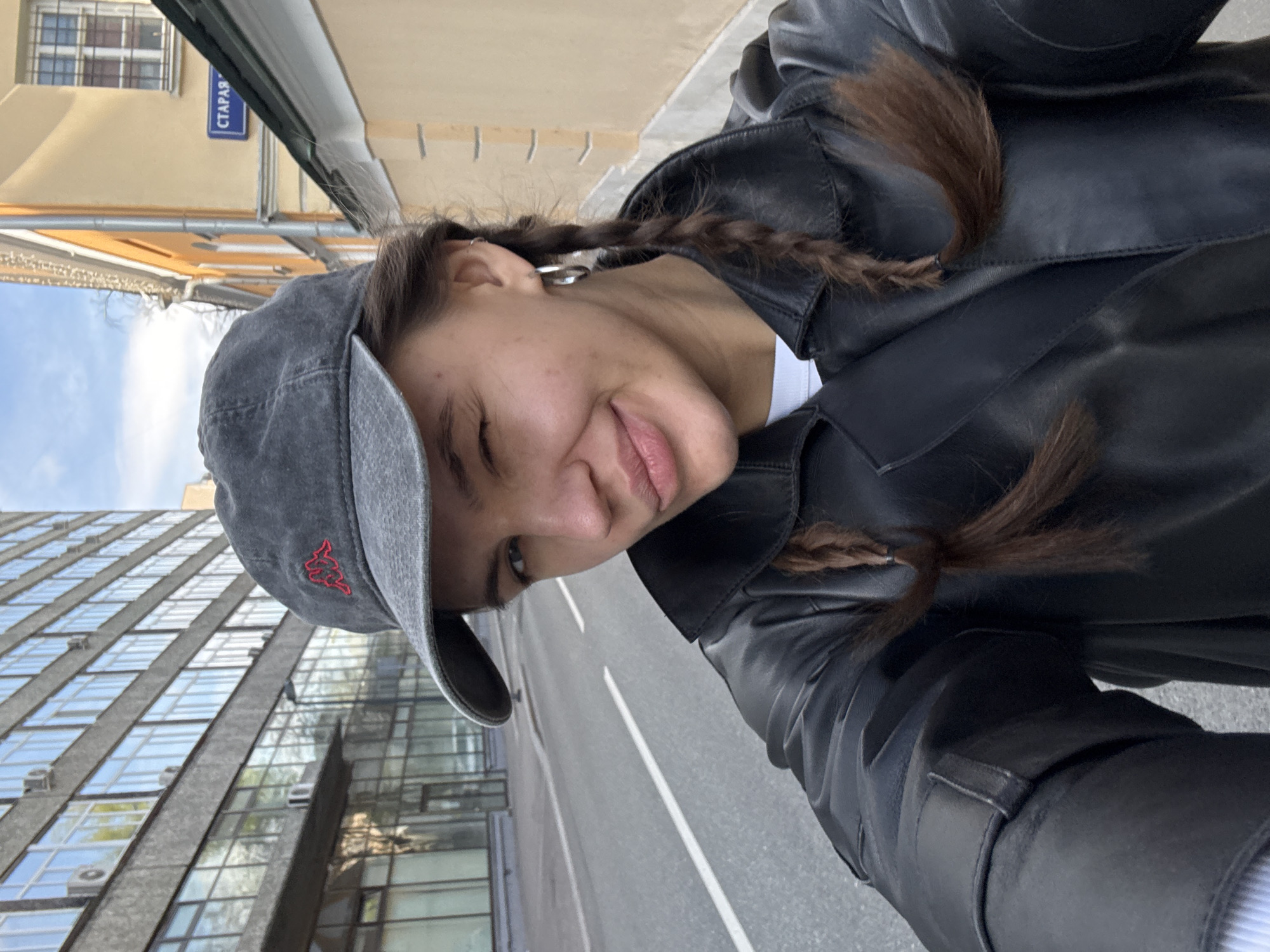 A woman with braided hair and a gray baseball cap poses for a selfie on a street in Moscow, Russia. She is wearing a black leather jacket. The street sign "СТАРАЯ" ("Old") is visible, suggesting the location might be a historic part of the city. A large, modern building with many windows is on the left side of the frame. The street is asphalt, and the sky is visible in the upper-left corner.