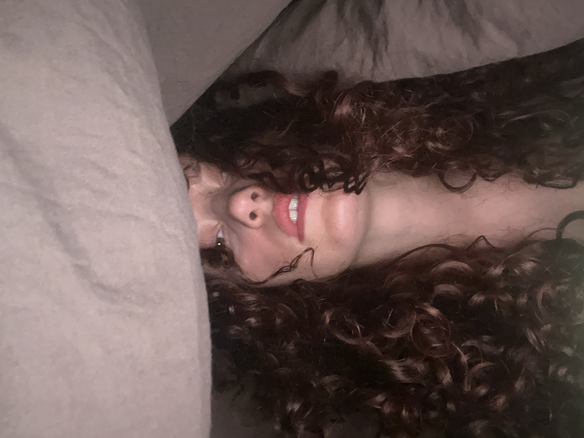 A woman lies in bed. Her head is tilted to the side, and she is partially covered by a beige blanket. She has curly, dark brown hair. Her lips are red, and she has a nose ring. No location information is included in the source image.