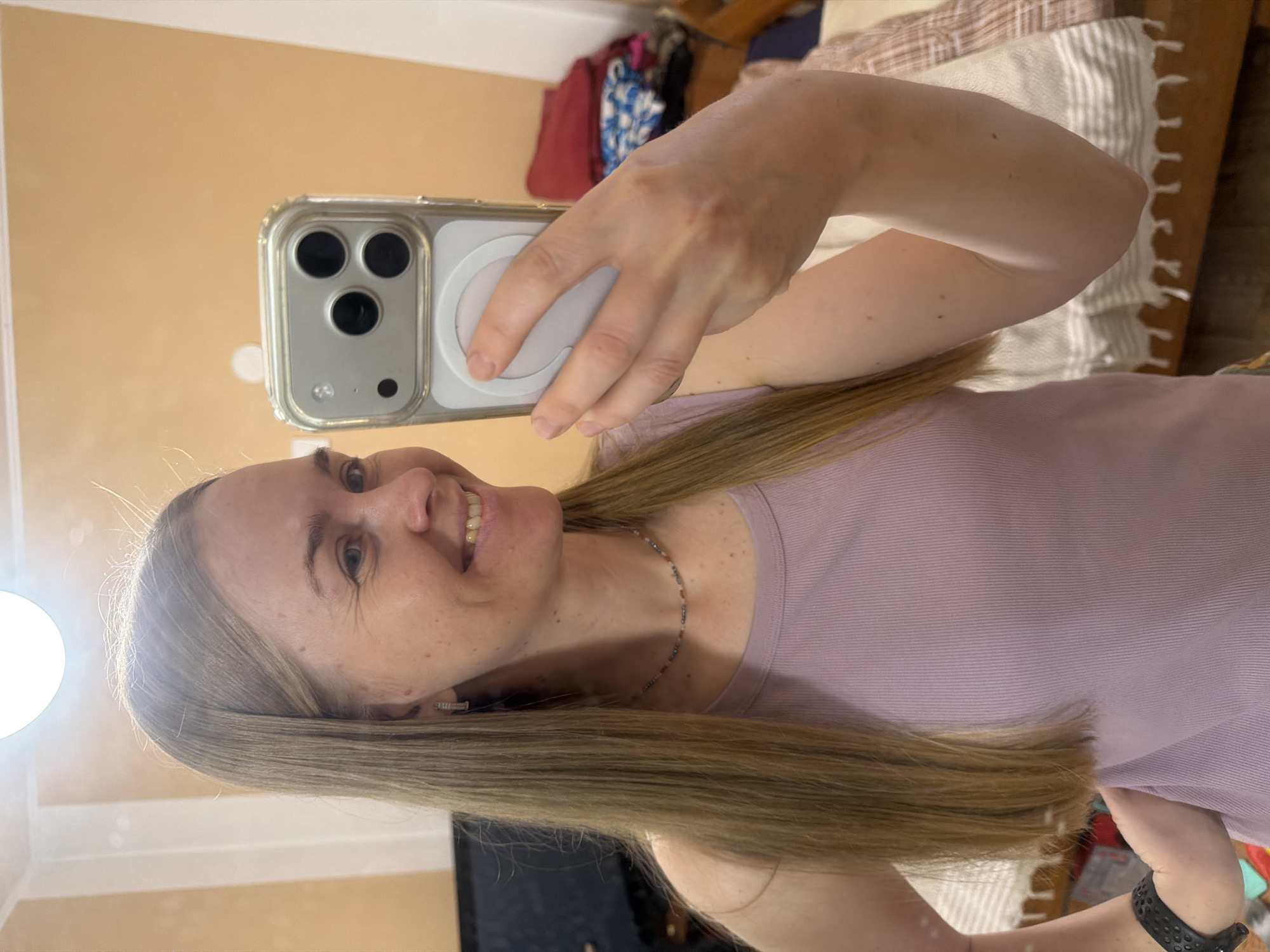 A woman with blonde hair is taking a mirror selfie in what appears to be a bedroom. She is wearing a light purple tank top and a beaded necklace. Her right hand is holding a phone with a silver case and a white circular accessory on the back. Her left arm is bent at the elbow, and she is wearing a black smartwatch. The background shows a wall painted a light orange-peach color, a white trim, and a bed with a patterned blanket. The overall lighting is bright, likely from an overhead light source. The image quality is good, with clear focus on the woman and her immediate surroundings. There are no discernable landmarks or indicators of the city of Khabarovsk, Russia, in the image.