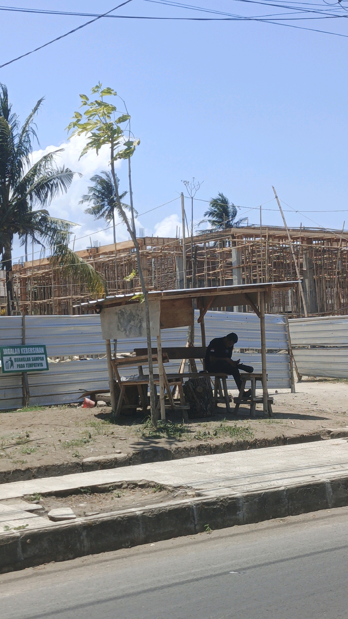 The image depicts an outdoor scene on a sunny day, likely in Pangandaran, Indonesia, given the presence of palm trees and the tropical weather. In the foreground, a damaged section of a sidewalk is visible, with debris and small plants growing in a large pothole. A person, dressed in dark clothing and a cap, is seated on a wooden bench beneath a makeshift shelter. The shelter appears to be constructed from salvaged materials and has a corrugated metal roof. The person is looking down at their hands, possibly using a mobile phone. Behind the shelter, a construction site is underway, with scaffolding and building materials visible. A white metal fence separates the construction area from the foreground. A green sign with Indonesian text, which translates to "Maintain Cleanliness, Throw Trash in its Place," is mounted on the fence to the left of the shelter. Various trees, including palm trees, are scattered throughout the scene. The sky is clear and blue with a few scattered clouds. The overall impression is one of a developing area with ongoing construction and everyday life continuing alongside it.