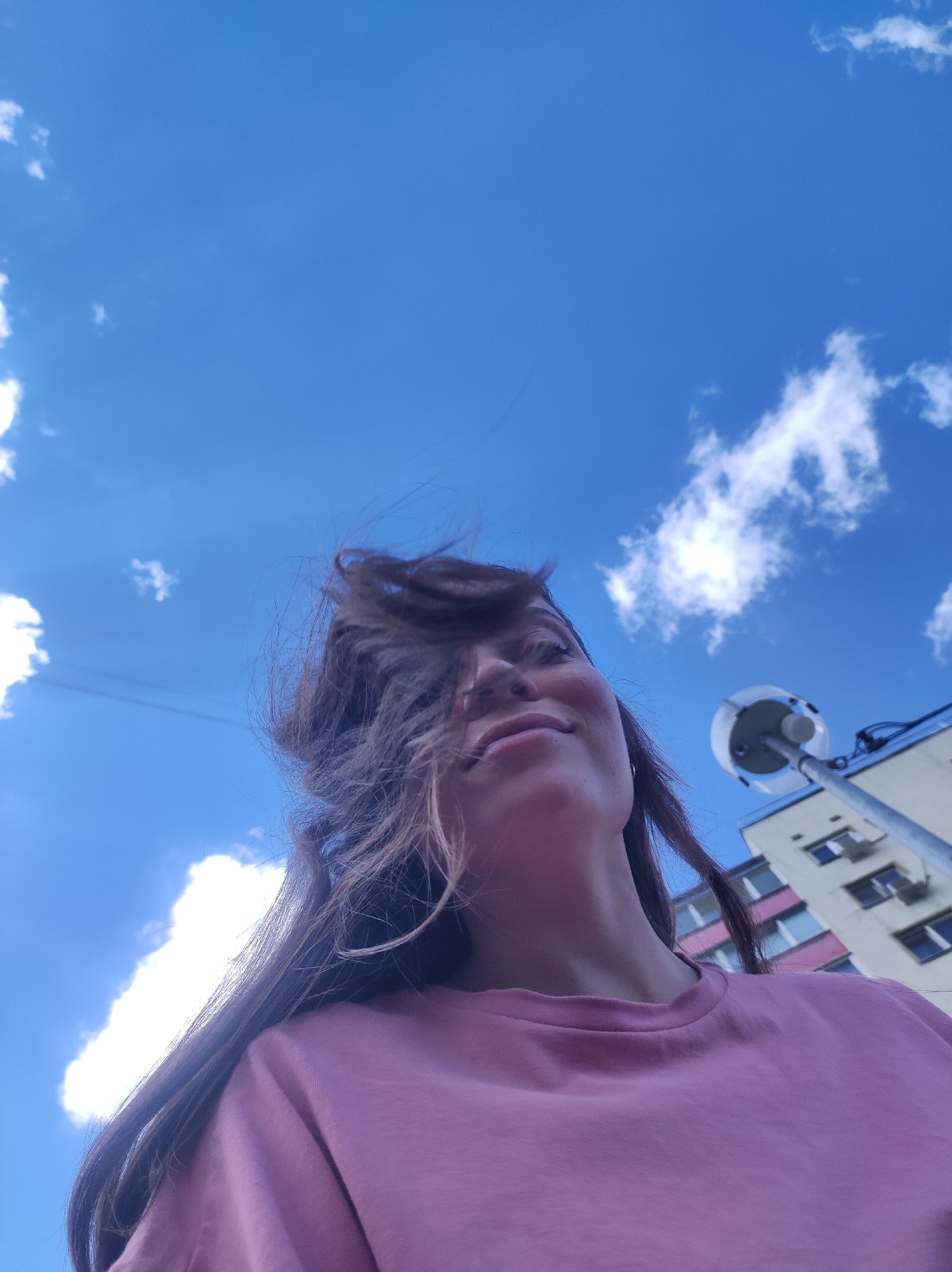 The image is a low-angle shot of a woman looking up at a blue sky with scattered clouds. Her brown hair is blowing in the wind, partially obscuring her face. She is wearing a pink t-shirt. In the background, a light-colored building with pink accents and several windows is visible, along with a street lamp. The scene appears to be outdoors in a city. Based on the provided context, the location is Chaikovsky, Russia.