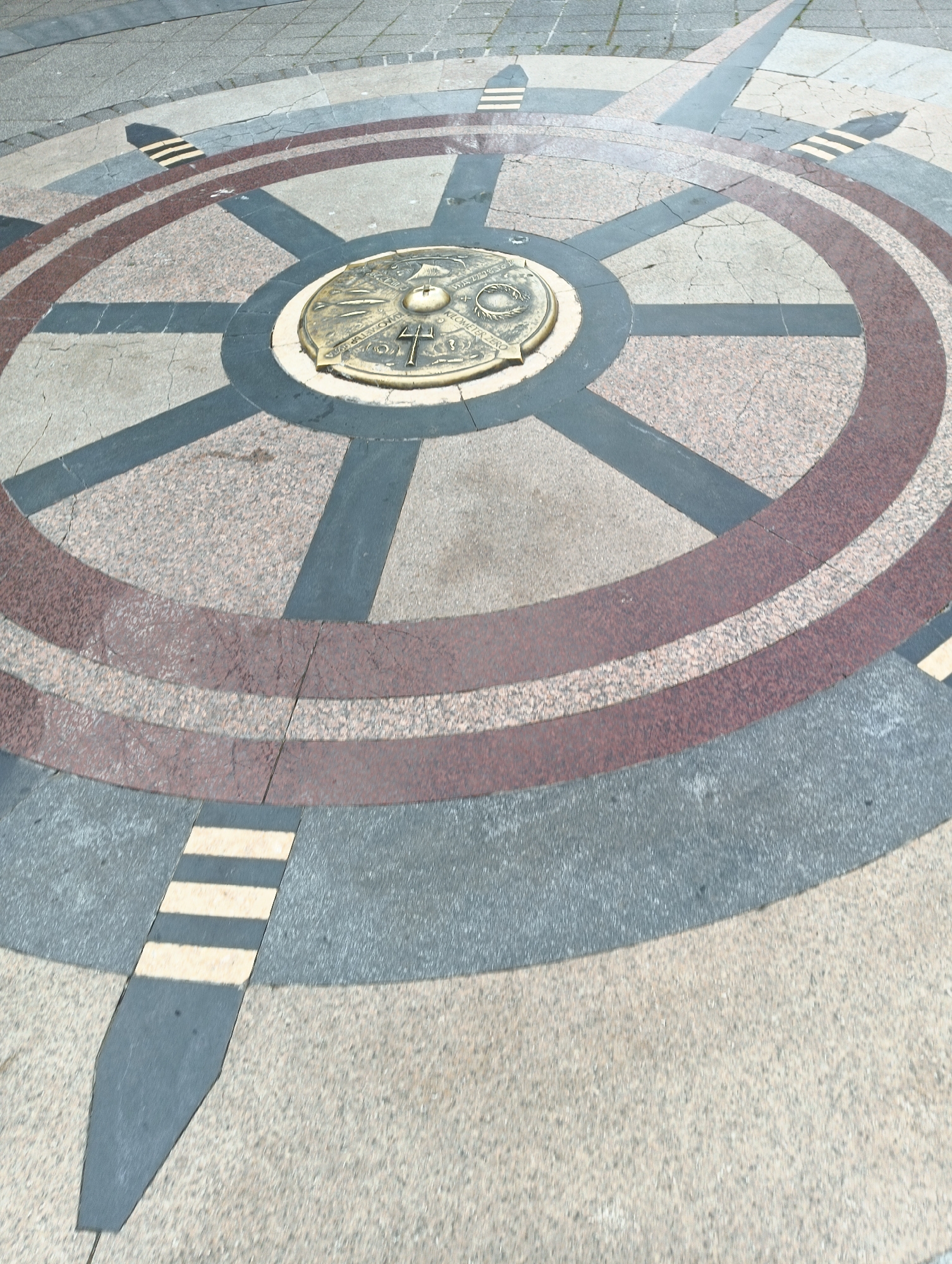 The image shows a decorative compass rose inlaid in a stone-paved surface. At the center of the compass rose is a bronze plaque. The compass rose design incorporates concentric circles and directional markers. The surface materials include various colors of stone and possibly granite. The ground appears to be outdoors. Based on the compass rose design, it is located in Burgas, Bulgaria.