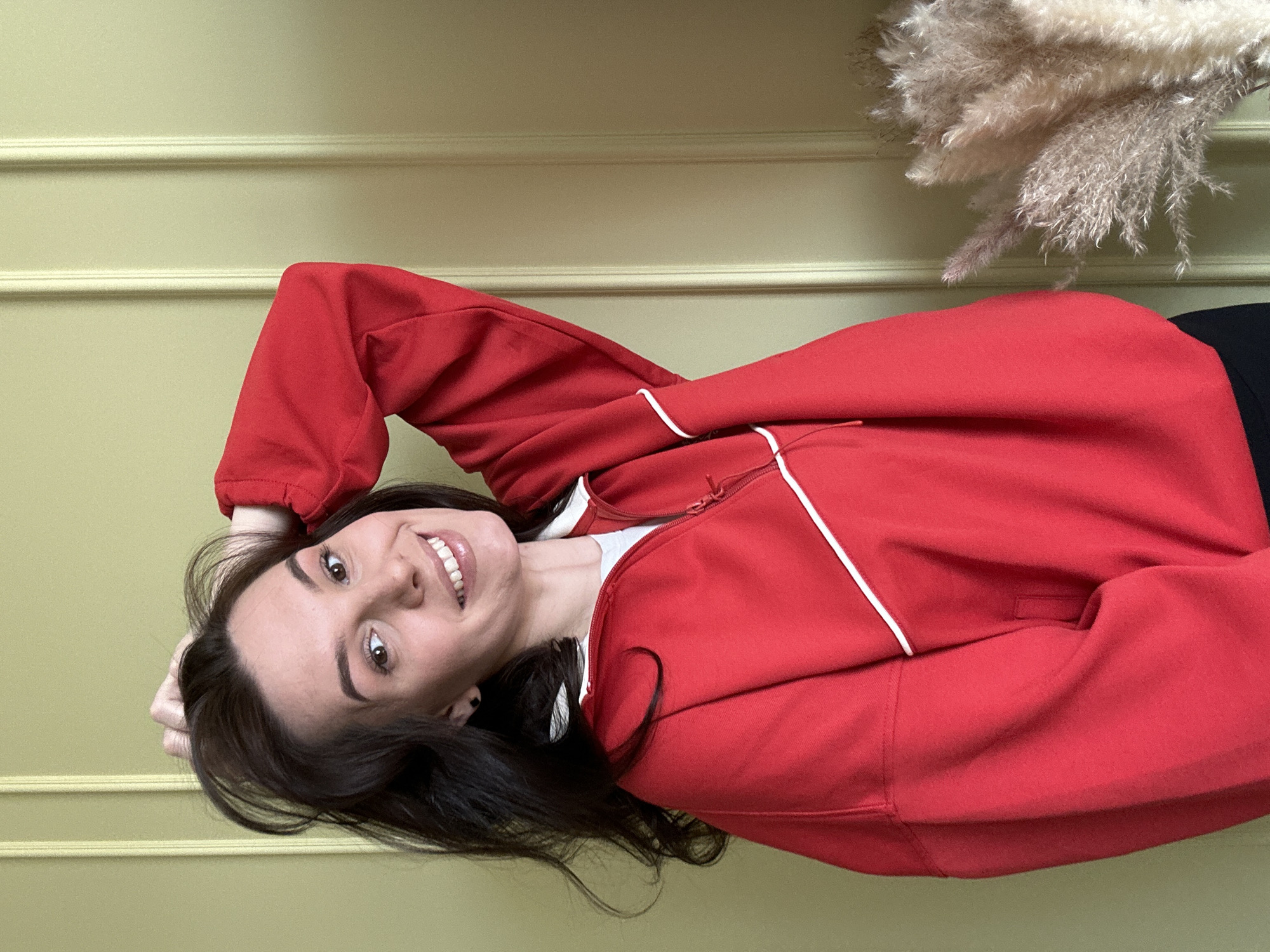 A woman with long dark hair is smiling and looking at the camera. She is wearing a red jacket with white piping and a white shirt underneath. She is resting her head on her hand. Behind her is a light green wall with decorative molding. To the right of the wall is a dried floral arrangement. The location is not evident and cannot be determined to be Chaikovsky, Russia.